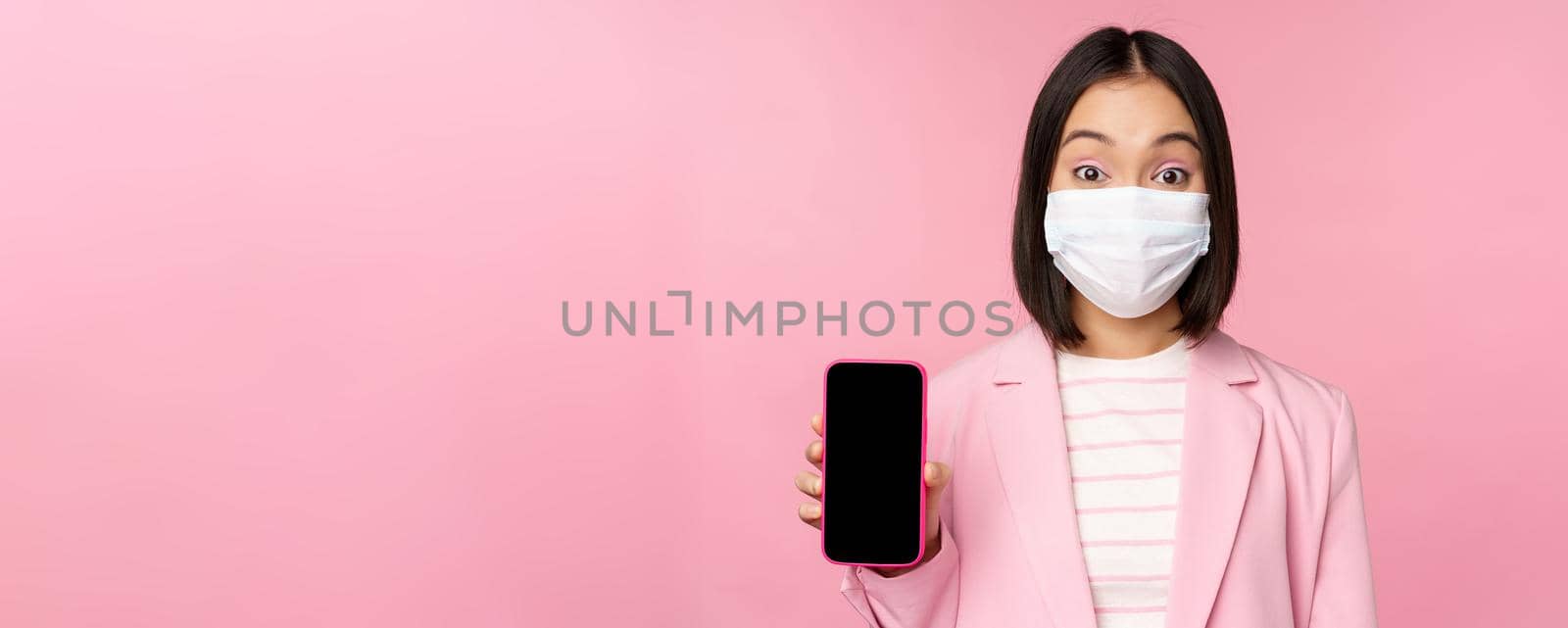 Portrait of smiling korean saleswoman in medical face mask, business suit, showing smartphone screen, standing over pink background by Benzoix