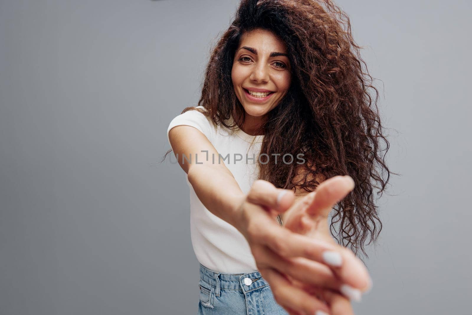 Wide angle view smiling happy Latin curly woman in white shirt summer casual clothes stretch hands to camera posing isolated at gray background. People emotion lifestyle concept. Copy space
