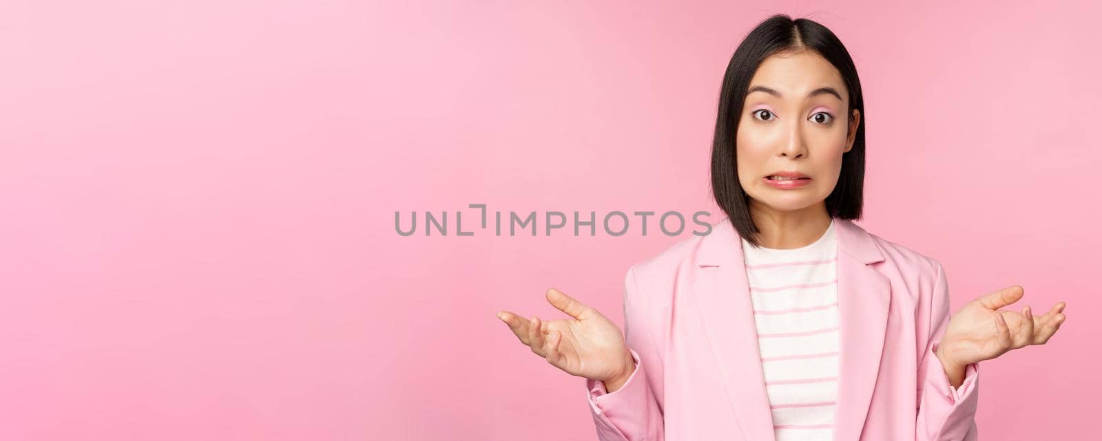 Portrait of confused asian businesswoman shrugging shoulders, looking clueless and puzzled, dont know, cant say, standing over pink background in office suit by Benzoix