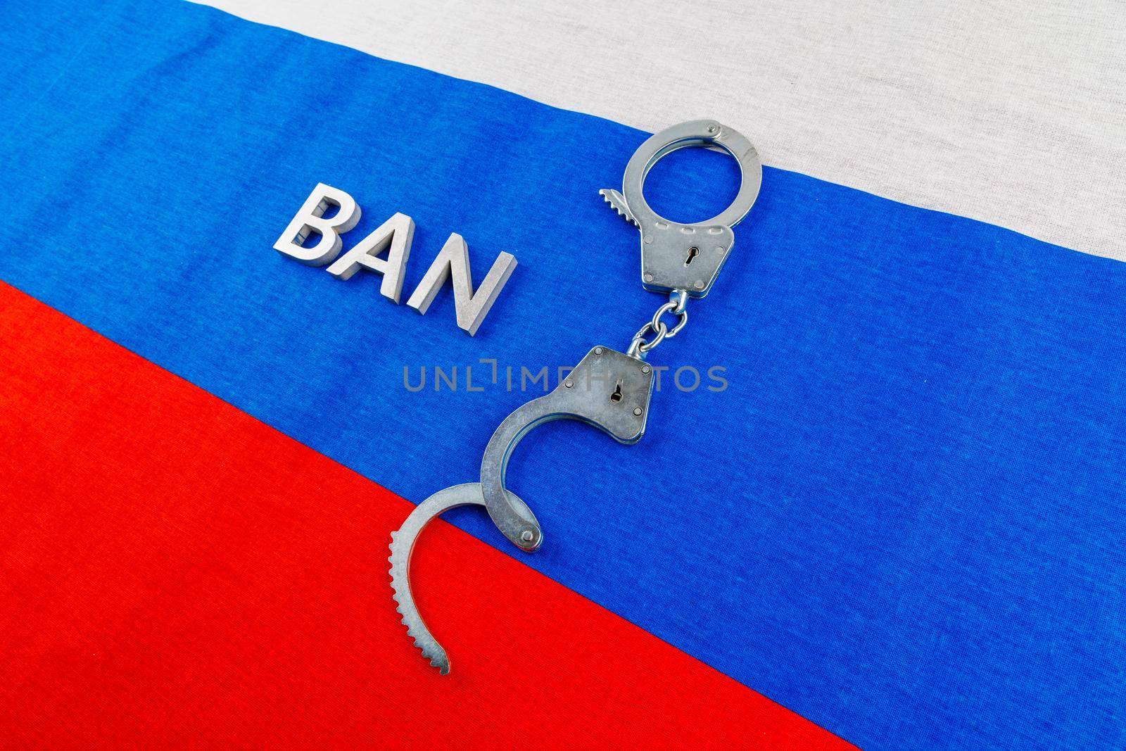 the word ban laid with silver metal letters near opened handcuffs over fabric tricolor flag of the Russian Federation