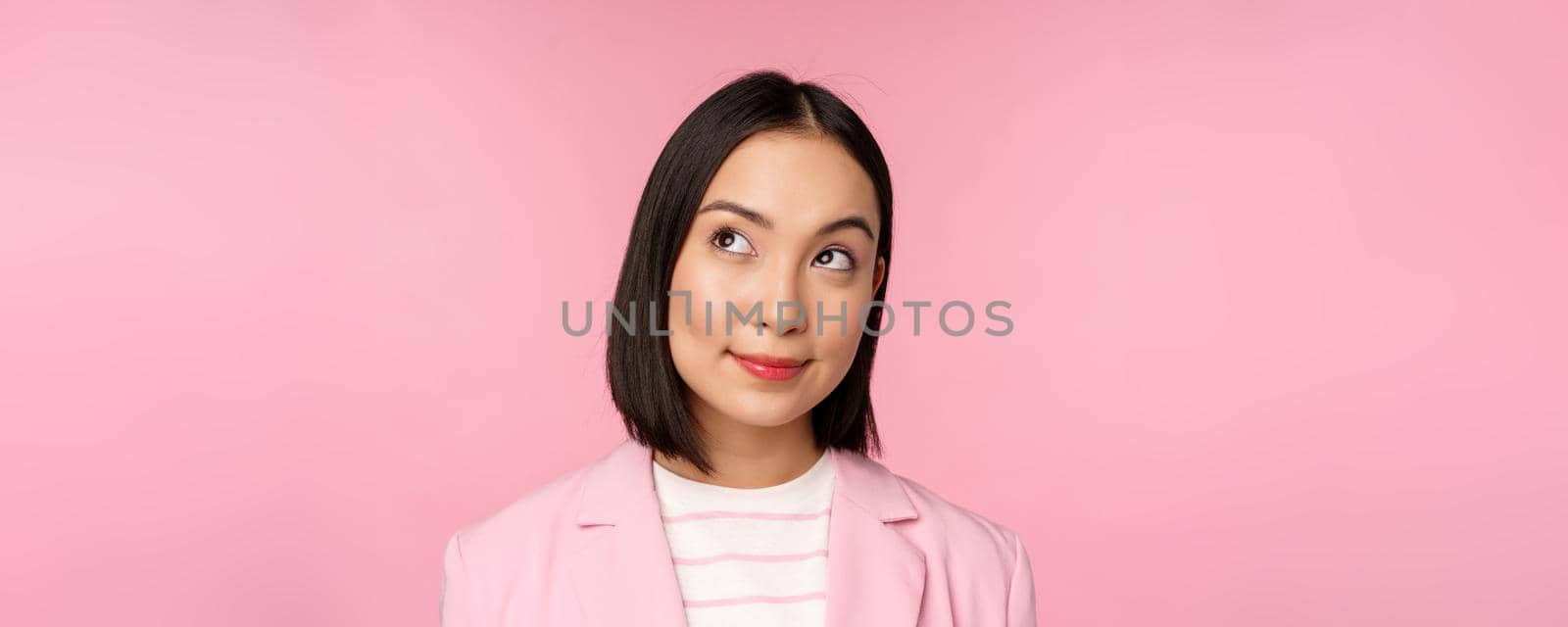 Close up portrait of young asian businesswoman thinking, smiling thoughtful and looking at upper left corner, standing over pink background.