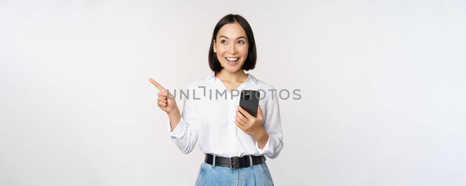 Excited asian girl shopping on mobile app, holding phone and pointing finger left at empty copy space, showing promo text, standing with smartphone against white background by Benzoix