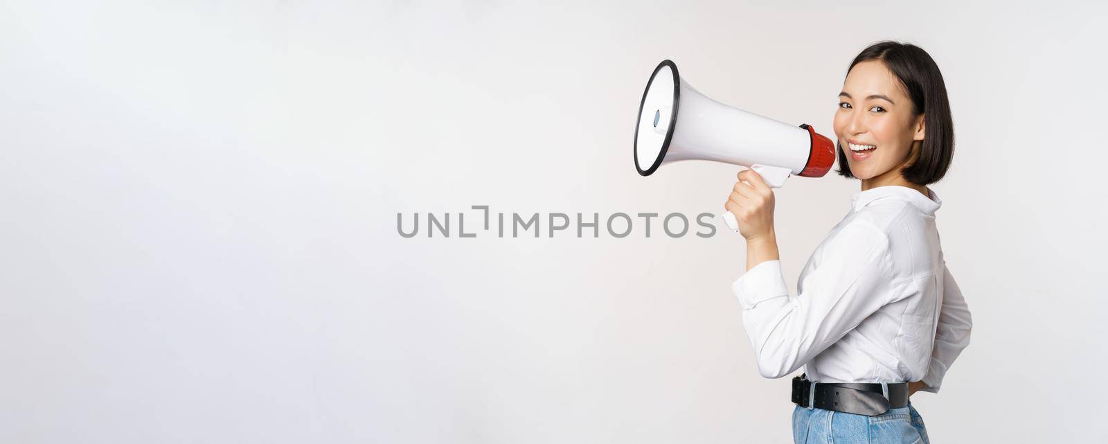 Beautiful young asian woman talking in megaphone, screams in speakerphone and smiling, making announcement, shout out information, standing over white background by Benzoix