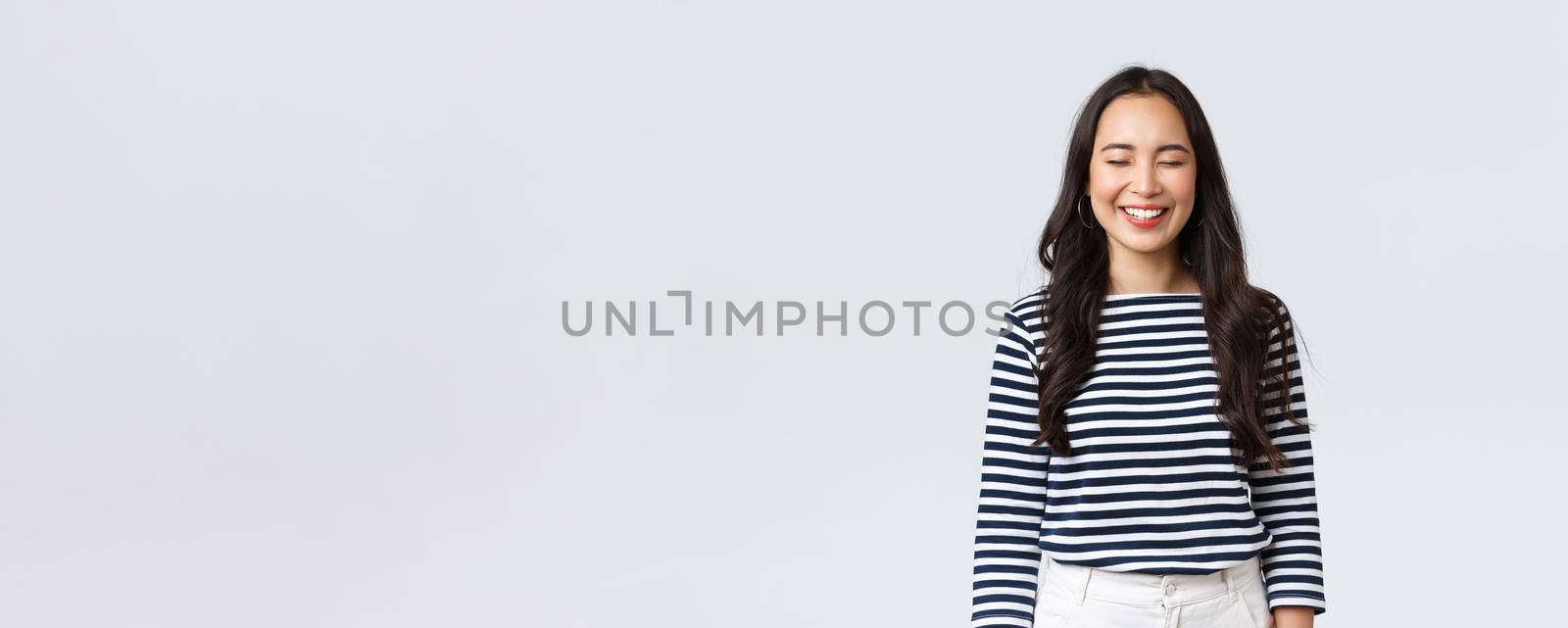 Lifestyle, people emotions and casual concept. Carefree, happy pretty asian woman in striped shirt close eyes and laughing sincere, having fun, funny moment, standing white background.