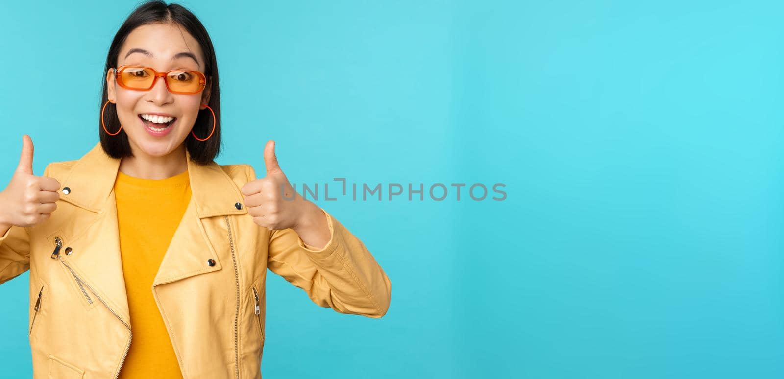 Enthusiastic korean girl looks excited and shows thumbs up, approves smth awesome, excellent choice and quality, stands over blue background.