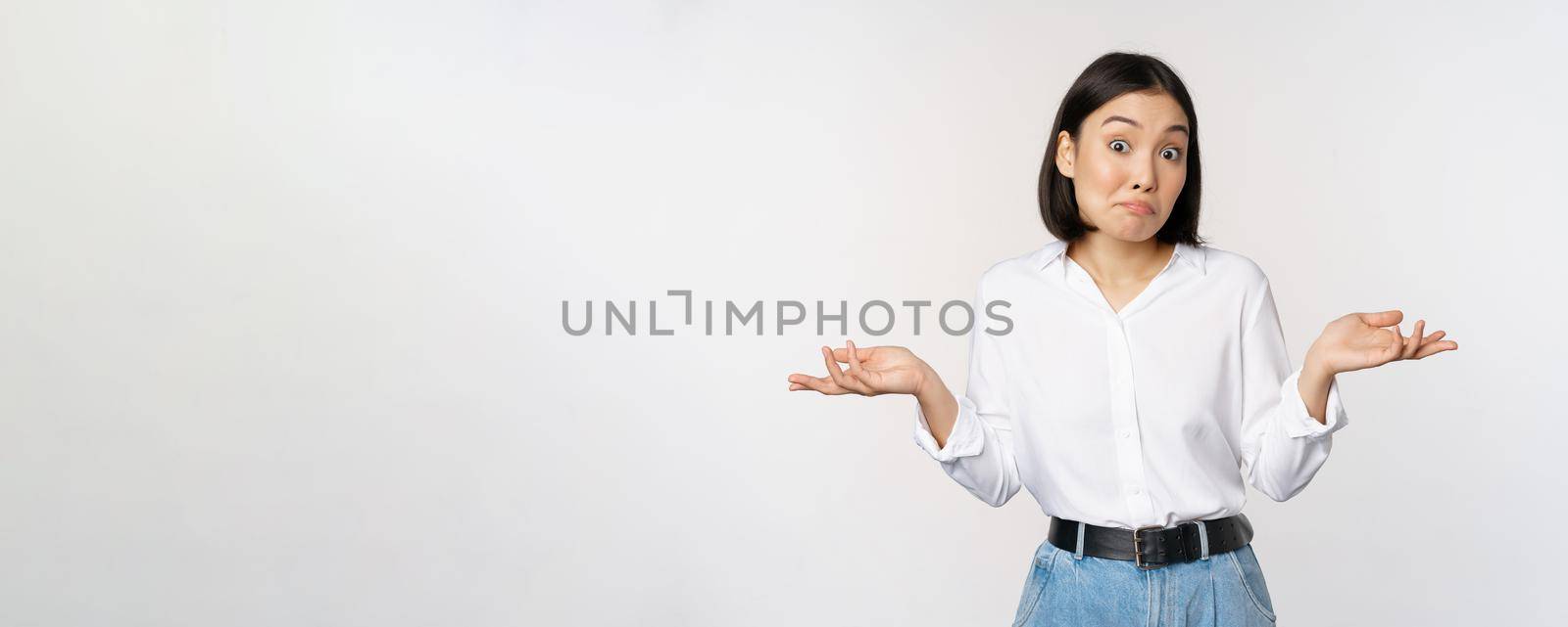 Image of confused asian office manager, office lady shrugging shoulders and looking clueless, standing puzzled against white background.