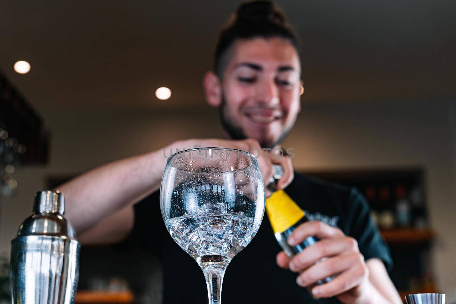 Young and modern bartender, with long dark hair, smiling, dressed in a black polo shirt, pouring liquor in a crystal glass to prepare a cocktail, at the wooden bar counter of the bar. Waiter preparing a cocktail. Cocktail glass with ice cubes. Gin Tonic. Bar filled with cocktail ingredients. Dark background and dramatic lighting. Low angle view