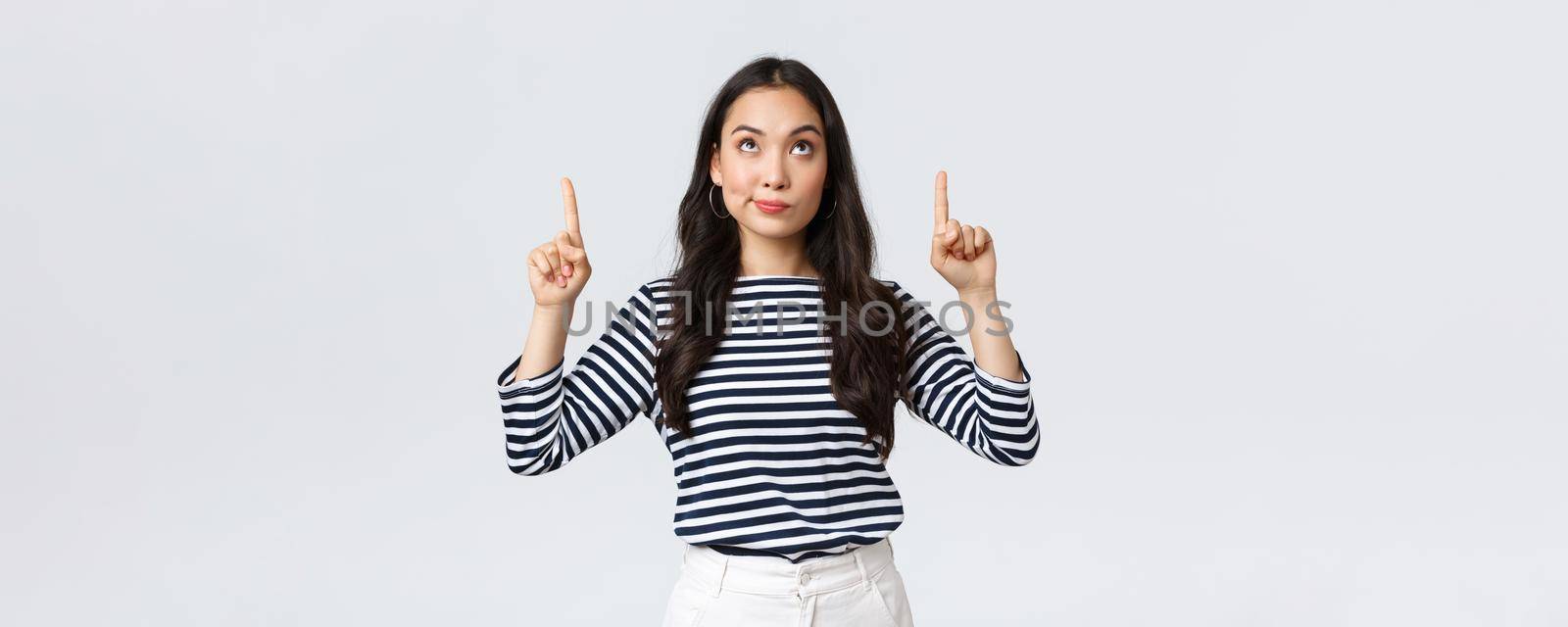 Lifestyle, beauty and fashion, people emotions concept. Indecisive and doubtful cute asian woman looking, pointing fingers up at strange promo, having doubts, hesitating before buying.