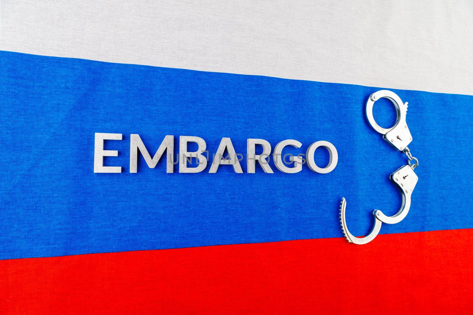 the word embargo laid with silver metal letters on russian tricolor flag with silver handcuffs in linear perspective