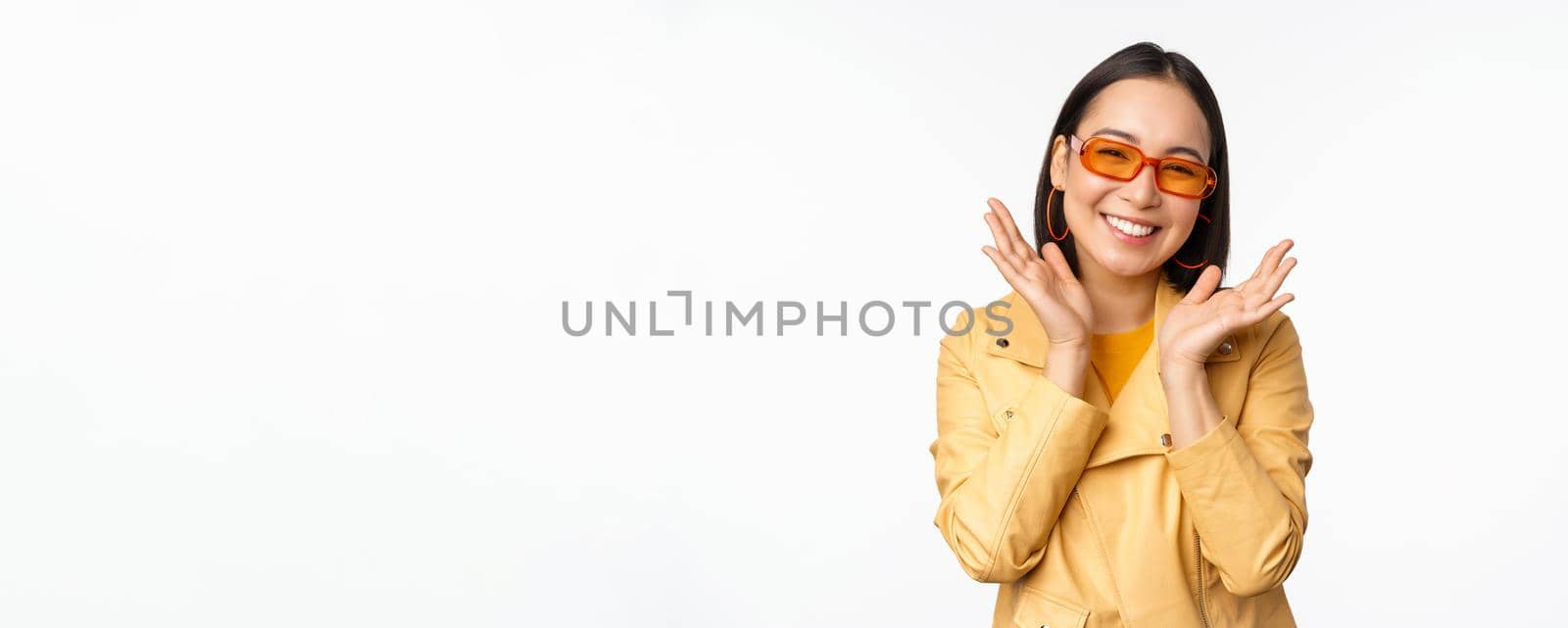 Beautiful asian girl in stylish sunglasses, smiling happy, looking bright and carefree, standing over white background. Copy space