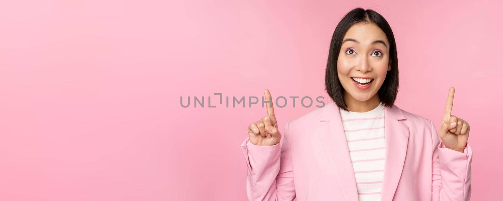 Enthusiastic corporate worker, asian business woman pointing fingers up and smiling, showing advertisement, logo, standing over pink background.