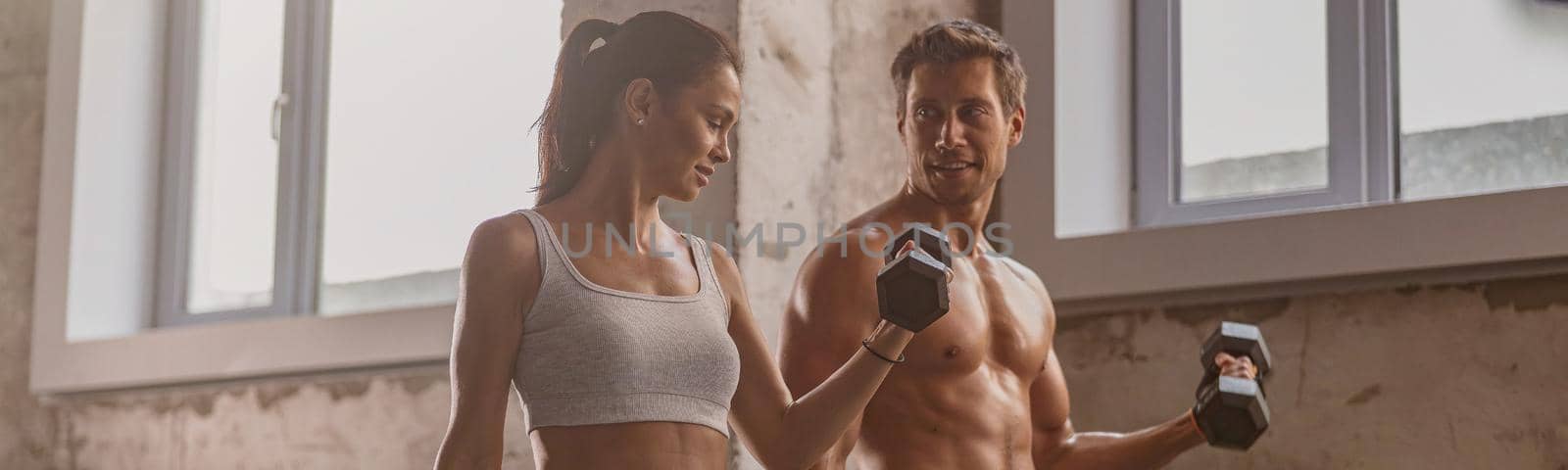 Young lady holding dumbbell and performing exercise for arms with strong athletic man in the gym