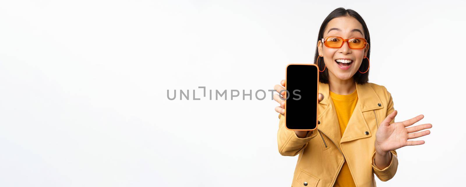 Enthusiastic asian female model, showing smartphone app interface, online store or website on mobile phone screen, standing over white background by Benzoix