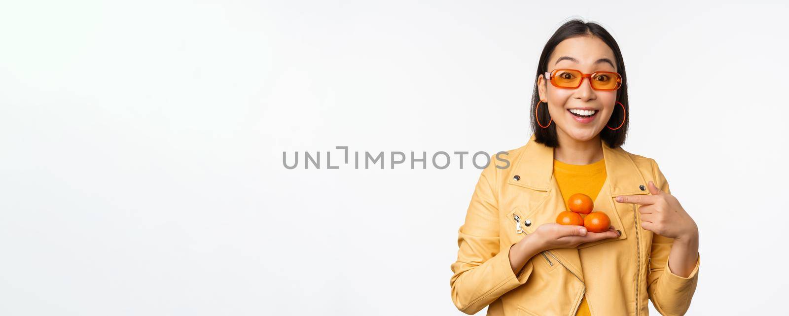 Stylish happy asian girl in sunglasses holding tangerines and smiling, posing against white background. Copy space