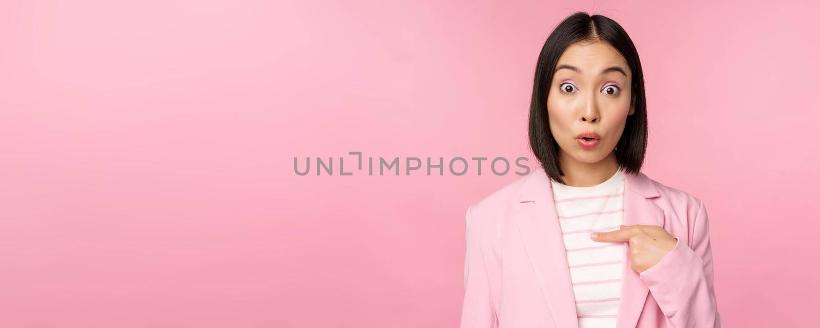 Portrait of asian businesswoman reacts surprised, points at herself with disbelief on face, posing in suit against pink background.