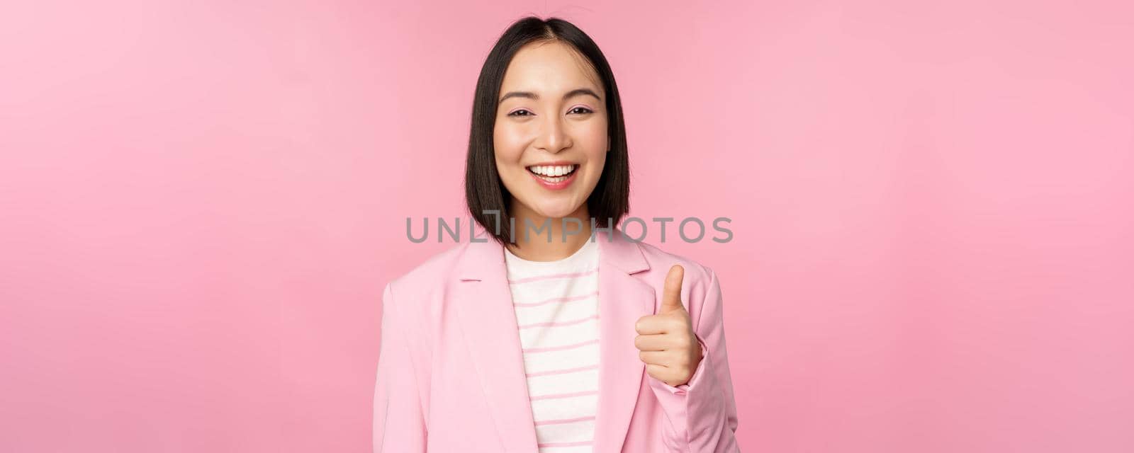 Portrait of asian businesswoman smiling satisfied, showing thumbs up, praise, like and approve, standing in suit over pink background by Benzoix