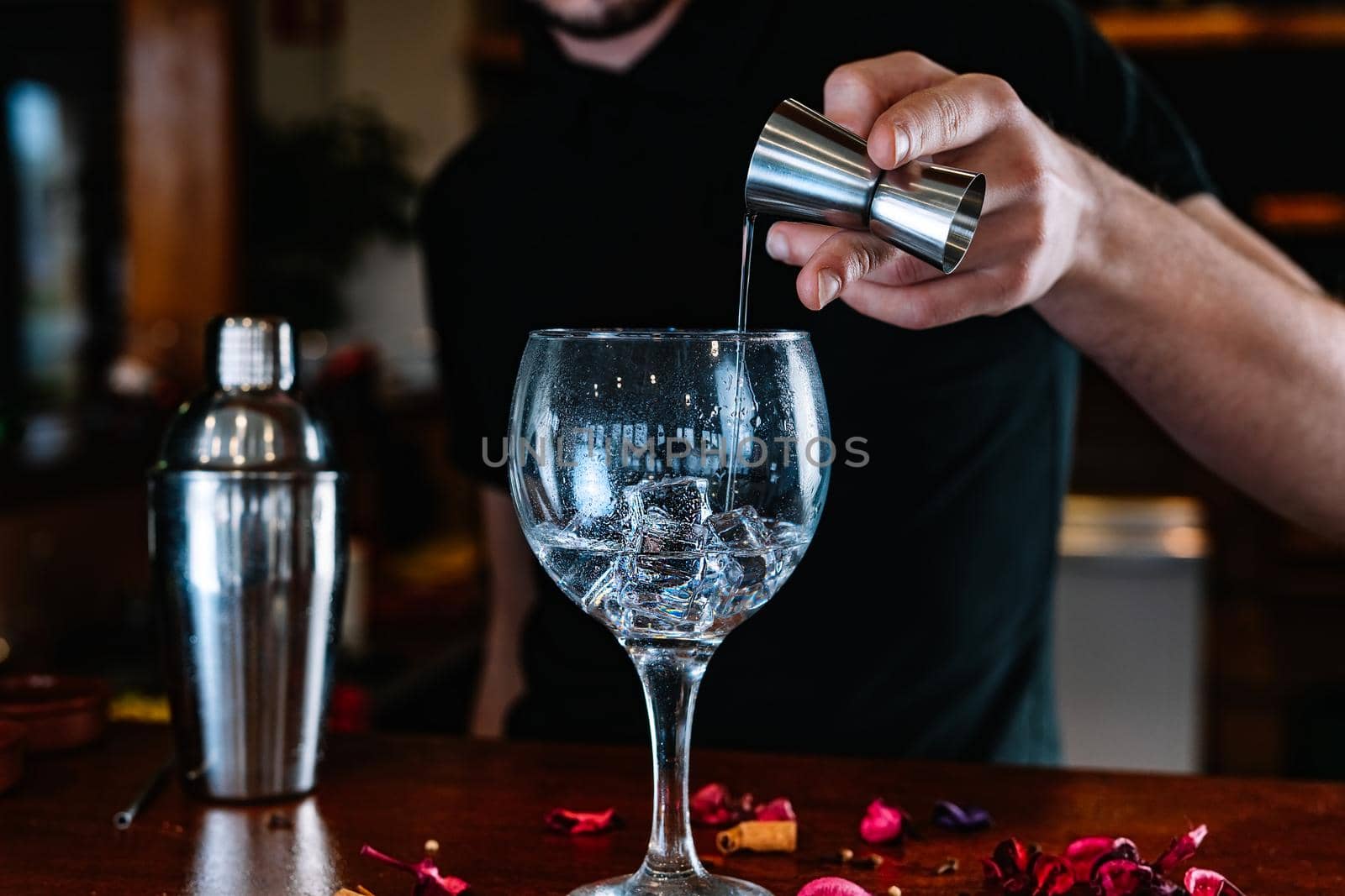 Young and modern bartender, with long dark hair, dressed in black polo shirt, pouring liquor into a crystal glass to prepare a mixed drink, on the wooden bar counter... Waiter preparing a cocktail. Cocktail glass with ice cubes. Gin Tonic. Bar full of cocktail ingredients. Dark background and dramatic lighting.