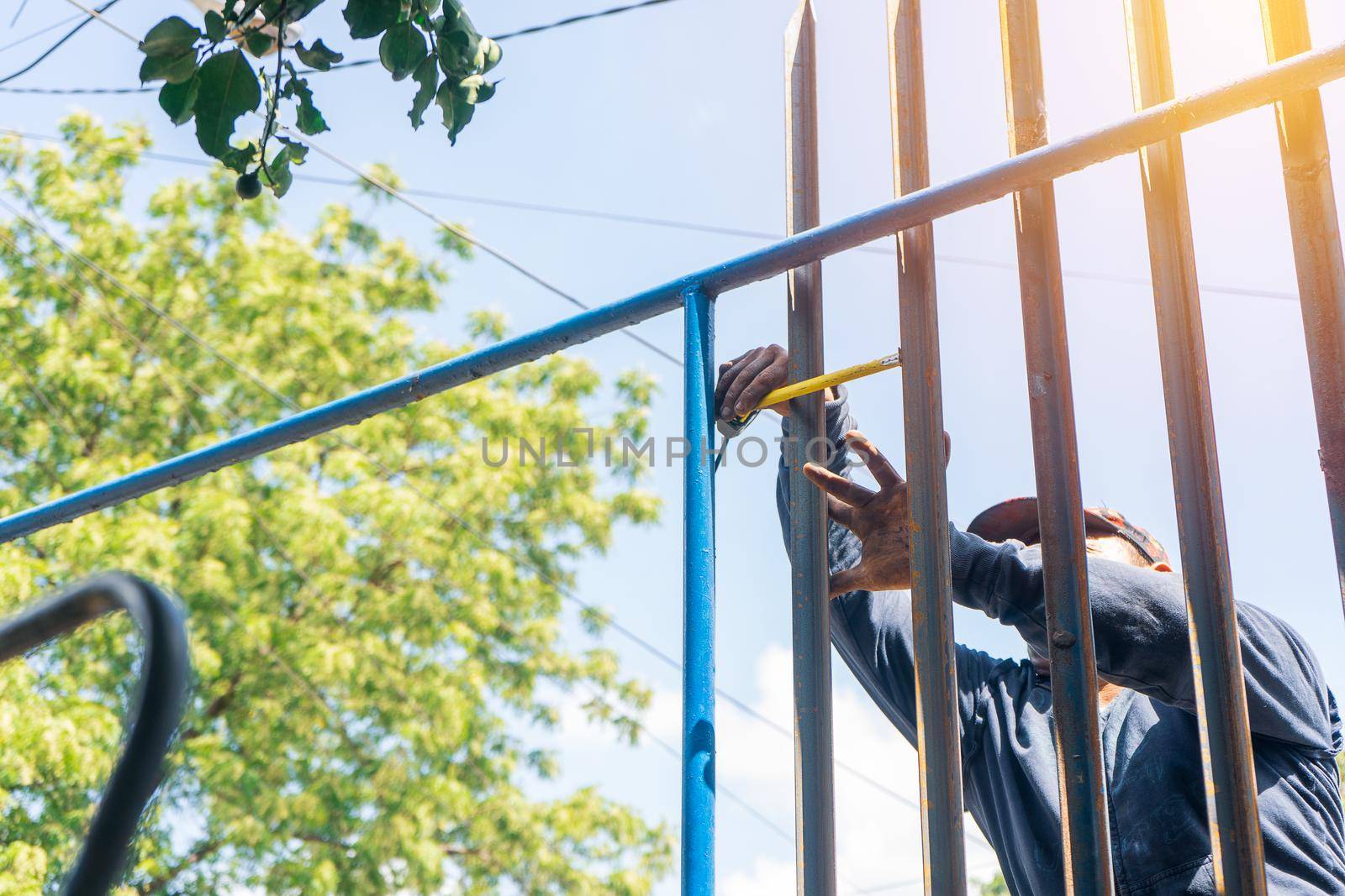 Blue collar worker outdoors measuring steel rods while installing a metal fence outdoors.