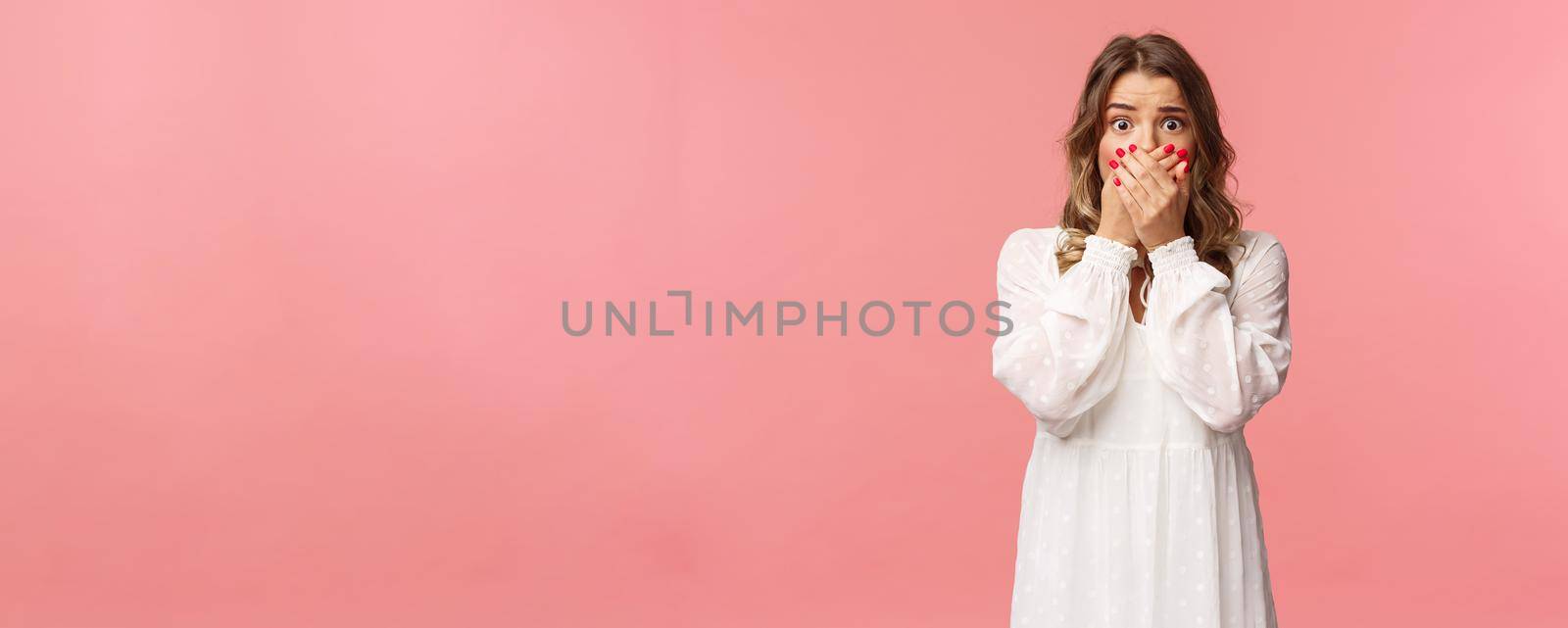 Shocked and anxious young concerned pretty girl in white dress, gasping close mouth with hands, look worried and startled, express fear or empathy with eyes, standing pink background.