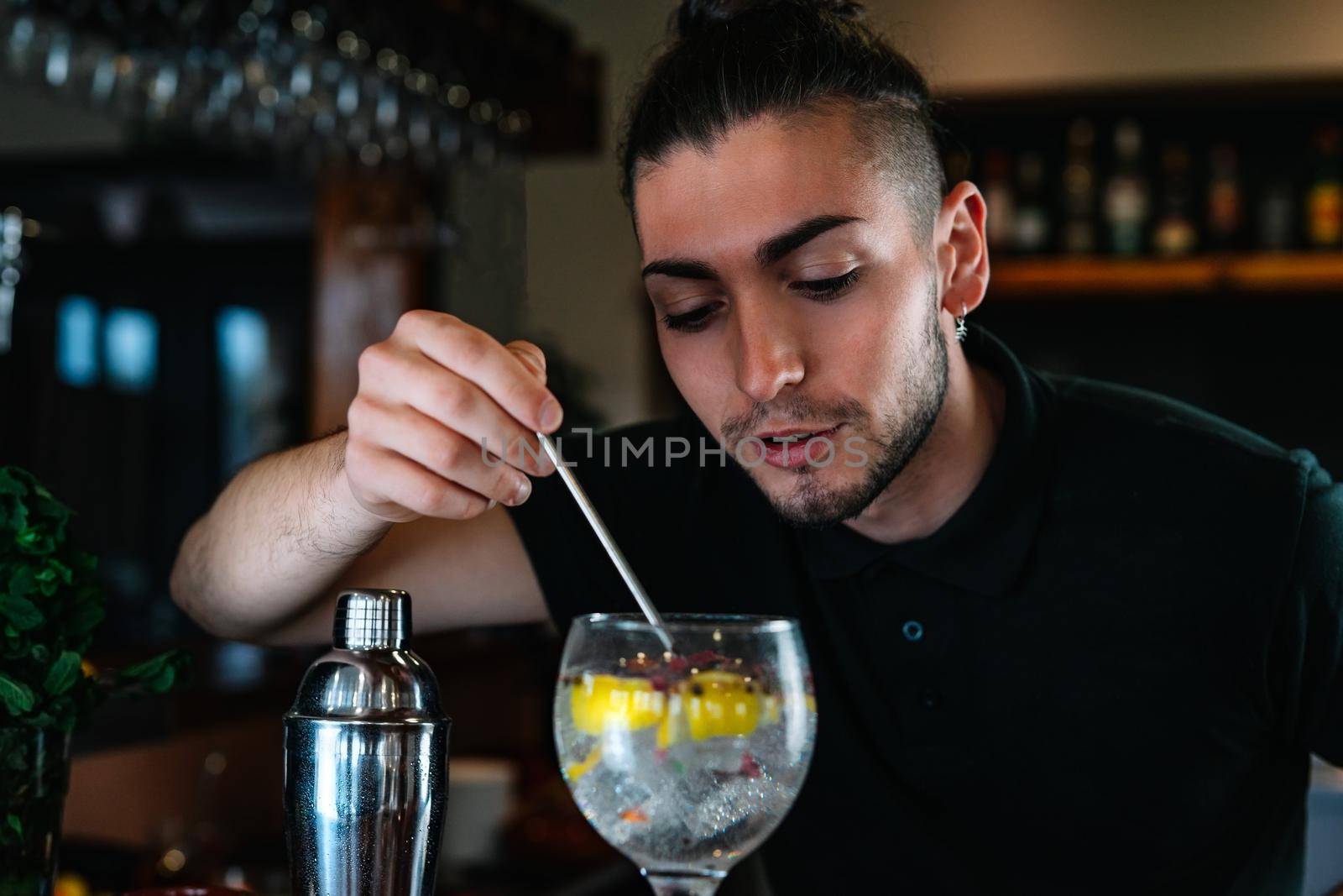 Young and modern bartender with long dark hair, dressed in black polo shirt, stirring a cocktail mix in a crystal glass with a slice of citrus fruit and ice for a cocktail. Waiter preparing a cocktail. Cocktail glass with ice cubes. Gin and tonic. Bar full of cocktail ingredients. Dark background and dramatic lighting.