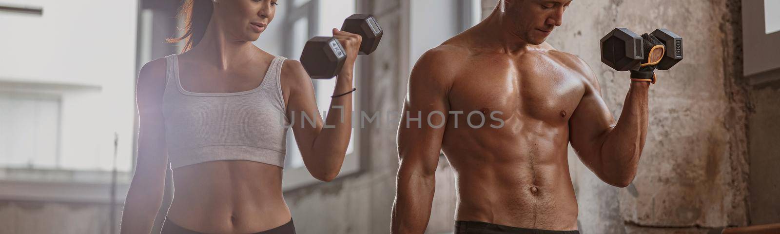 Slender young female and athletic man holding black dumbbells and doing hand exercise together in the gym