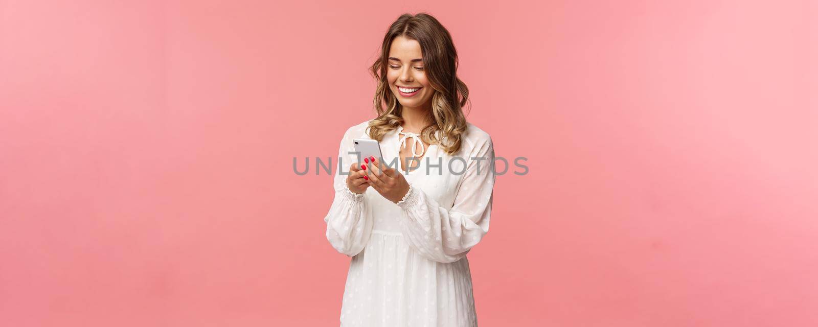 Technology and smartphones concept. Attractive young woman in cute white dress messaging, arrange romantic date, booking tickets via online application on mobile phone, smiling at display.