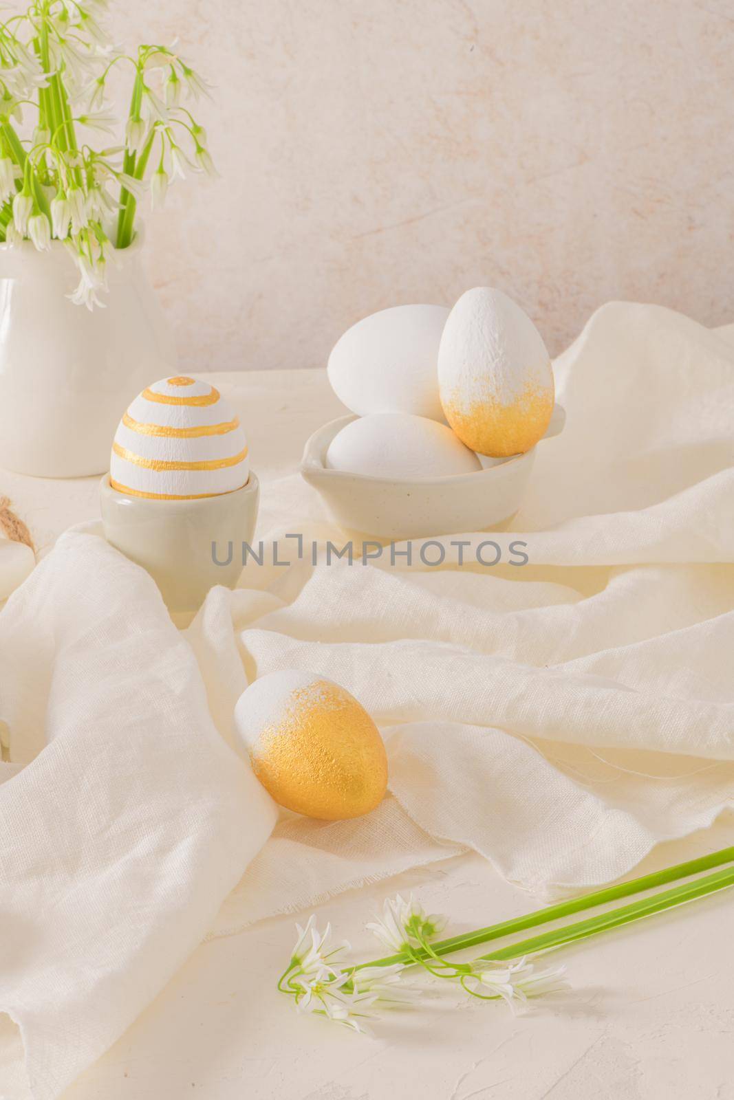 Close up of golden and white Easter eggs with white flower on bright white wooden table background.