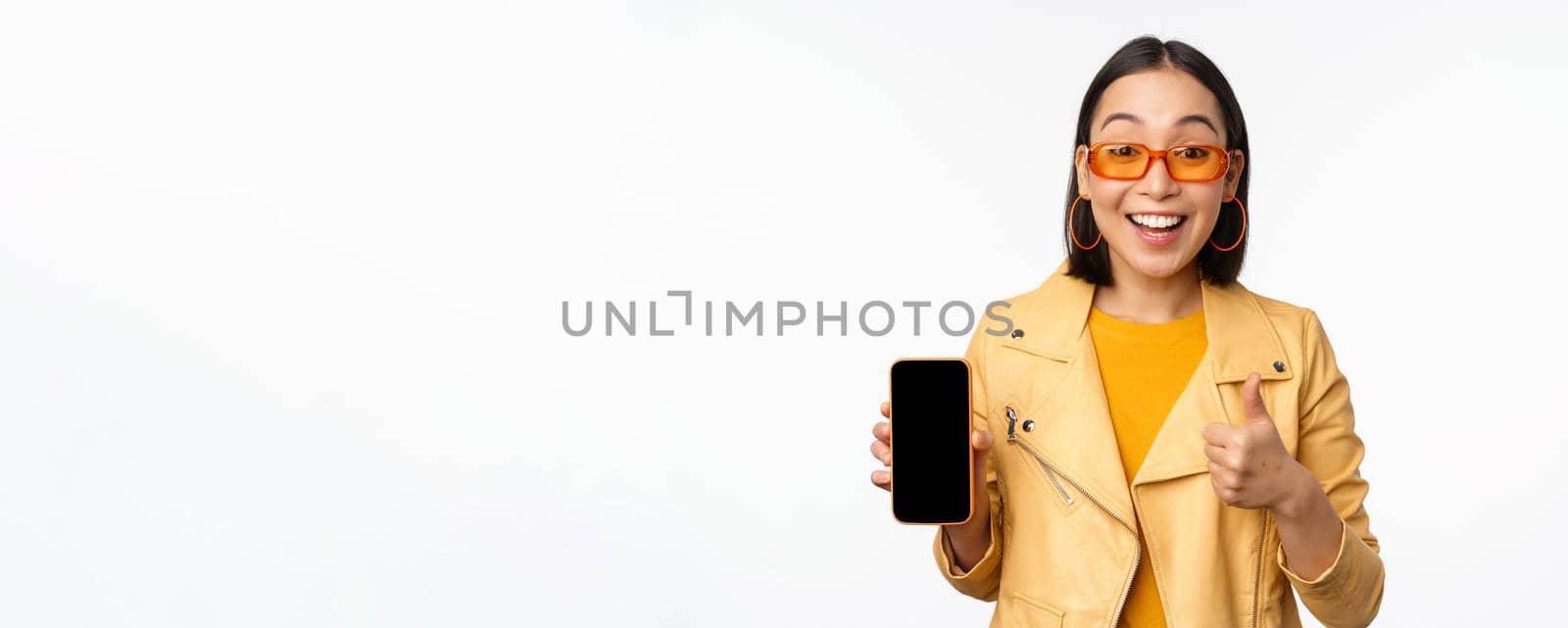 Beautiful korean girl, asian woman in sunglasses, showing smartphone app interface, thumbs up, recommending mobile phone application, white background.