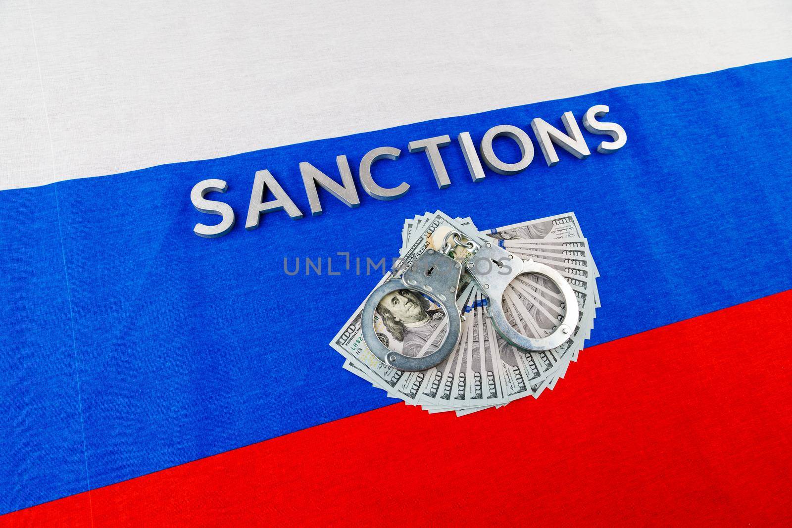 the word sanctions laid with silver metal letters on russian tricolor flag near dollar banknotes and handcuffs over it in linear diagonal perspective