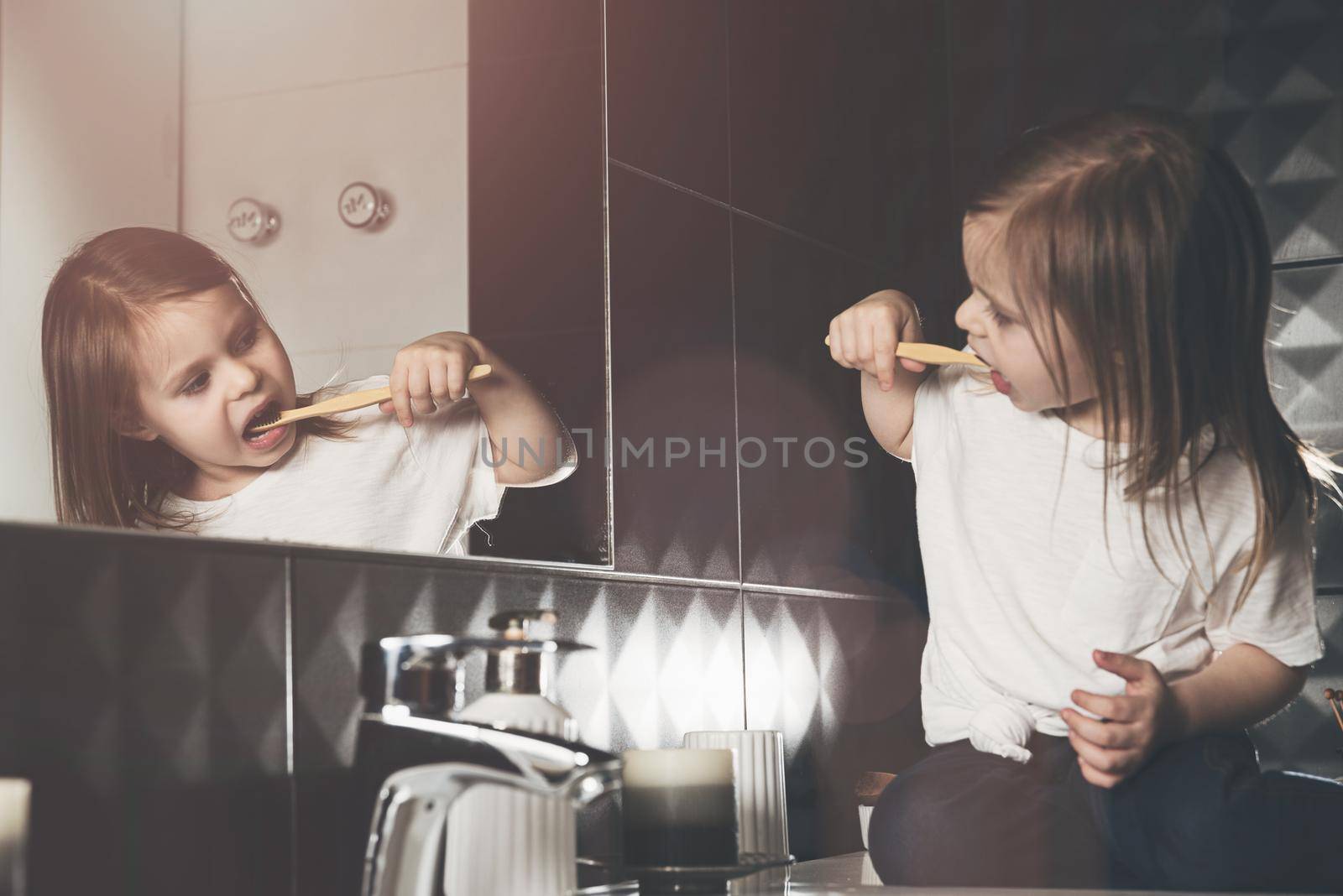 A small child in blue jeanse and white t-shirt brushes his teeth with a bamboo toothbrush. eco friendly. Black tiles on a wall. White sink