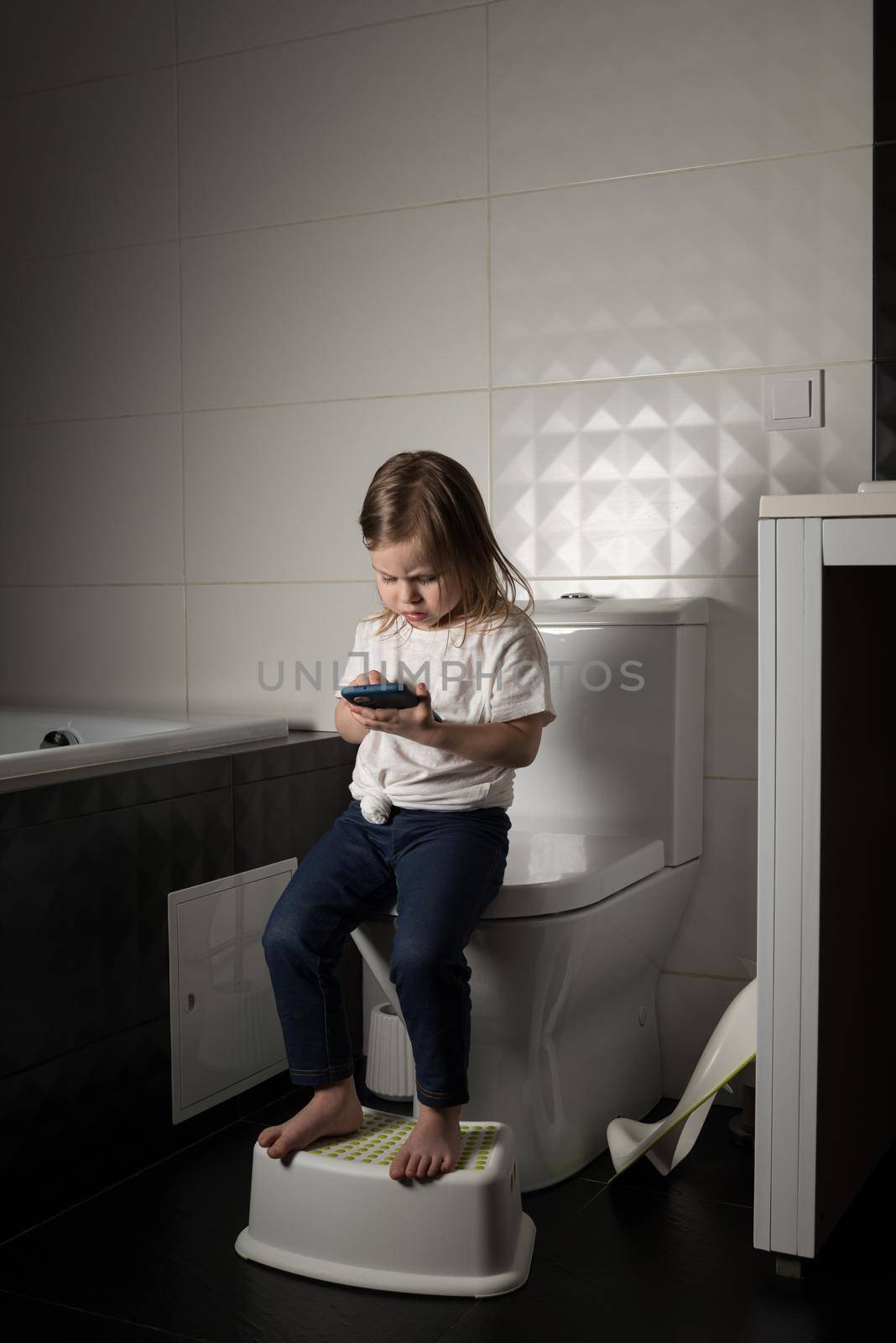 A girl dressed in a blue jeance and white t-shirt playing with a mobile phone in the bathroom by Ashtray25