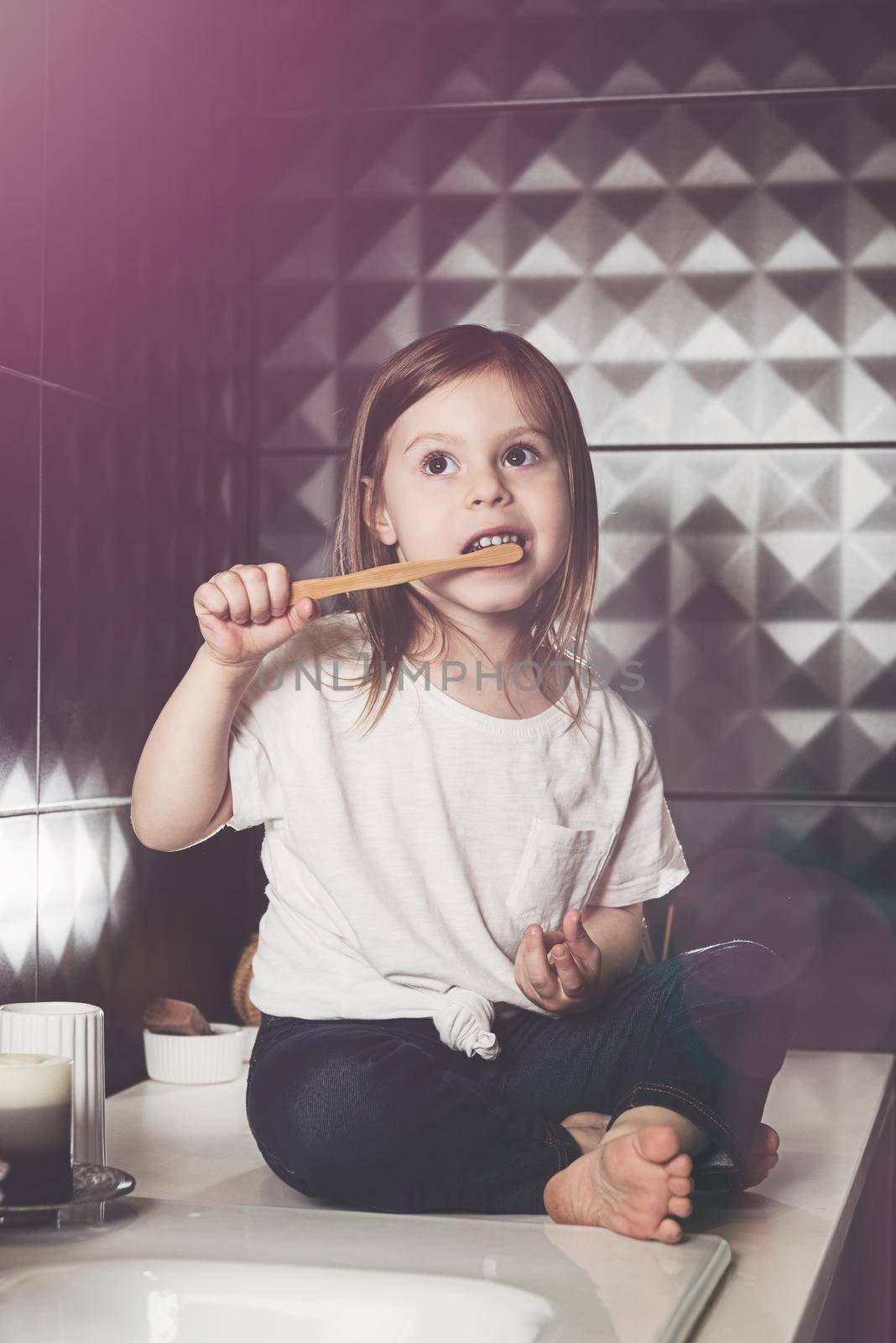 A small child in blue jeanse and white t-shirt brushes his teeth with a bamboo toothbrush. eco friendly. Black tiles on a wall by Ashtray25