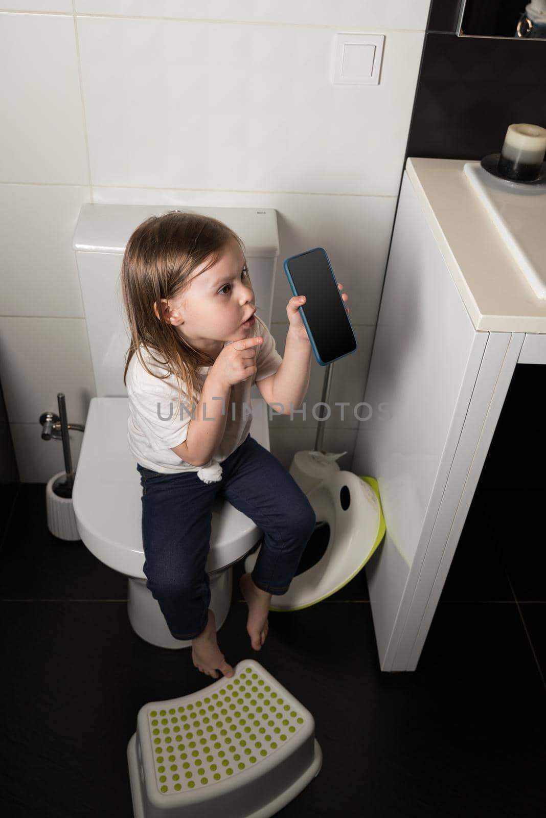 A girl dressed in a blue jeance and white t-shirt playing with a mobile phone in the bathroom.