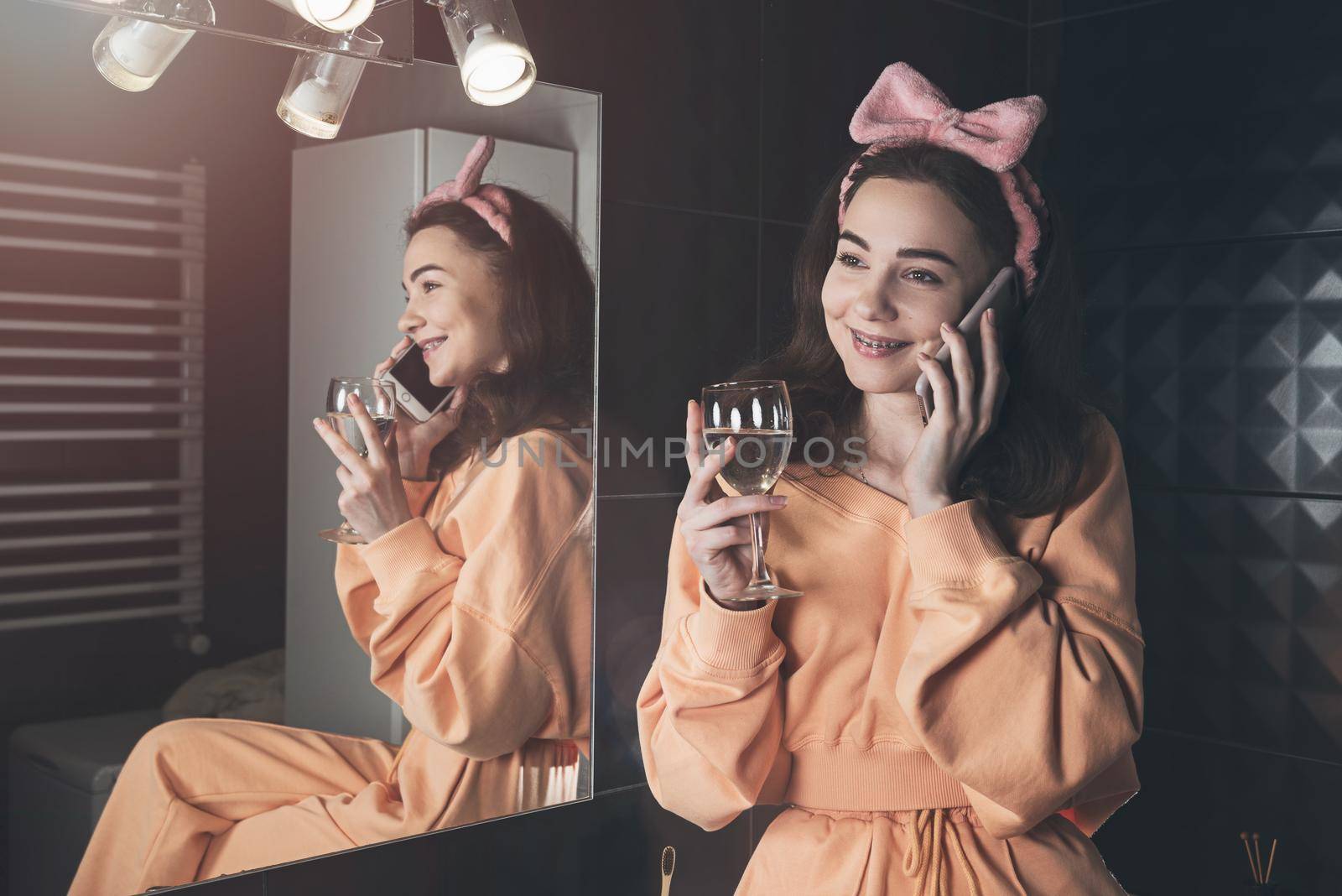 Pretty funny woman with mobile phone in the bathroom whit a glass of water in a hand. Woman wearing orange pajamas and a bow on his head