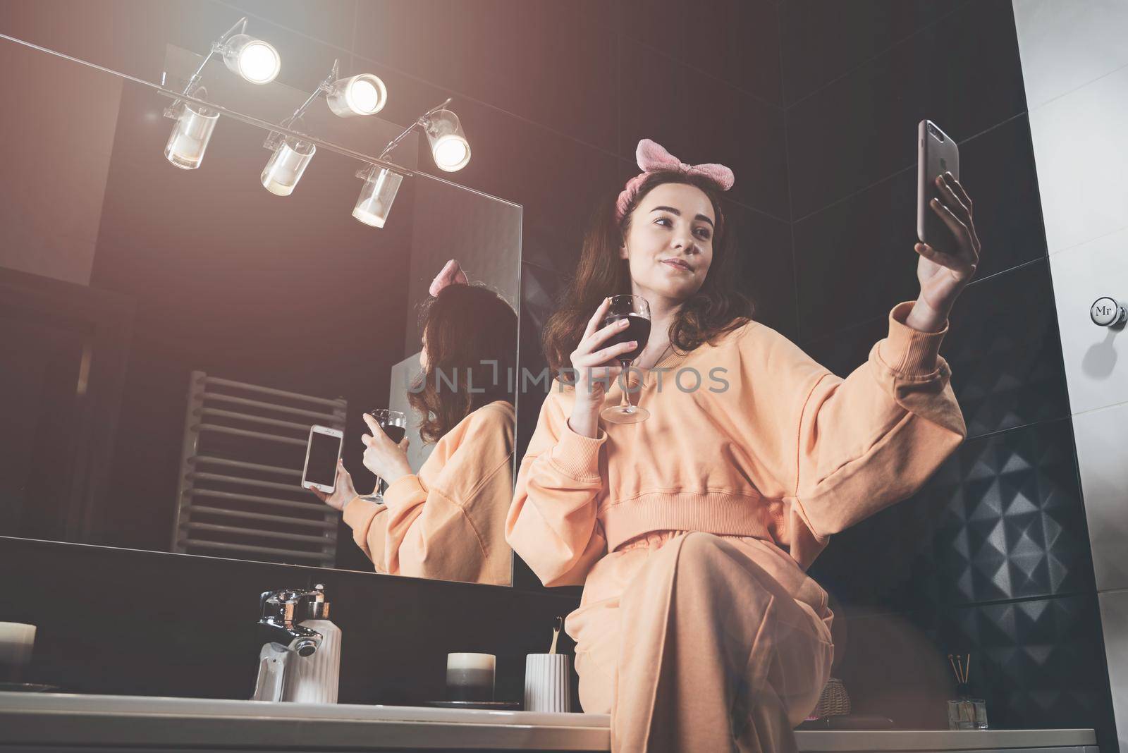 Attractive young woman in a bathroom communicating on a smartphone and drinking wine. Relaxation and leisure concept. girl dressed in orange pajamas