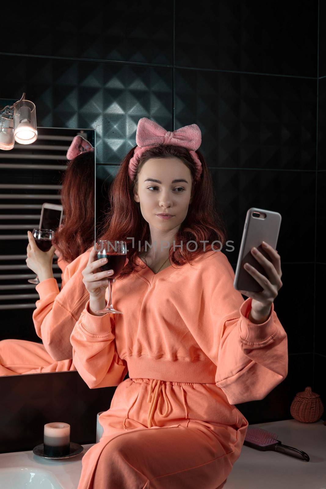 Attractive young woman in a bathroom communicating on a smartphone and drinking wine. Relaxation and leisure concept. girl wearing in orange pajamas