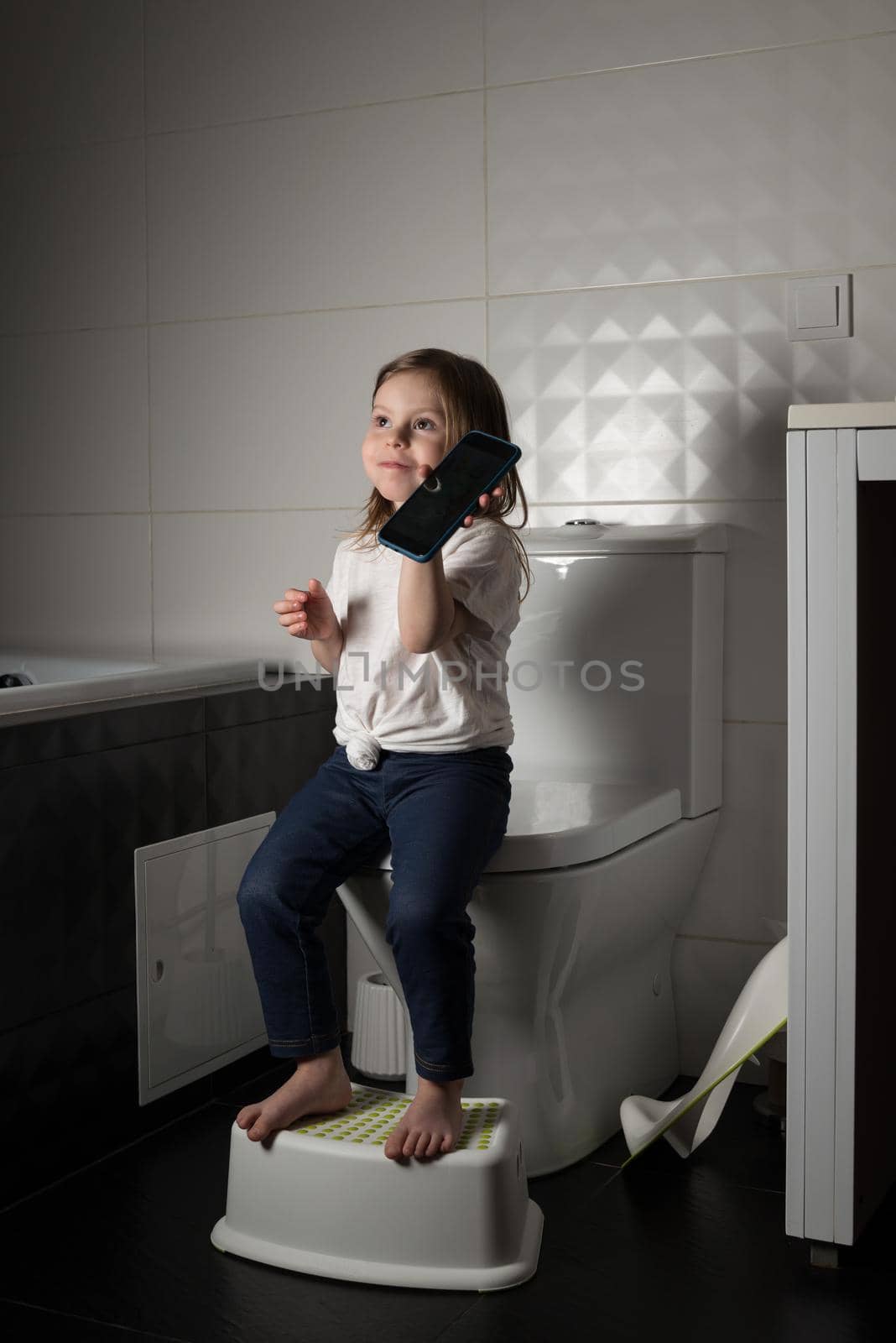 A girl dressed in a blue jeance and white t-shirt playing with a mobile phone in the bathroom.