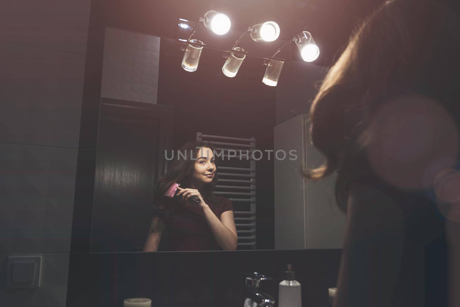 Young woman brushing hair in front of a bathroom mirror. Black tiles on a wall