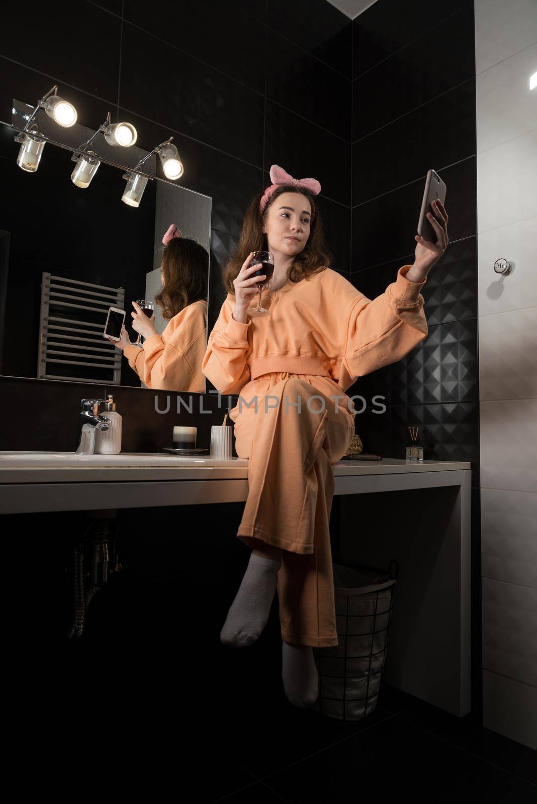 Attractive young woman in a bathroom communicating on a smartphone and drinking wine. Relaxation and leisure concept. girl wearing in orange pajamas