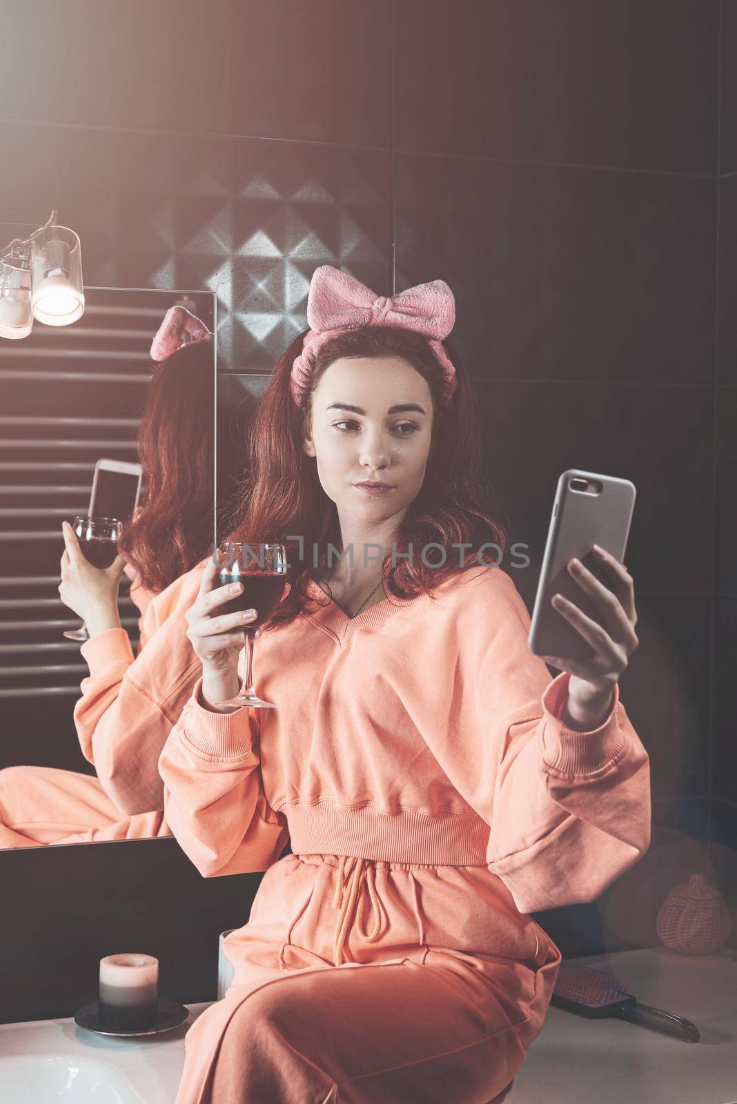 Attractive young woman in a bathroom communicating on a smartphone and drinking wine. Relaxation and leisure concept. girl wearing in orange pajamas