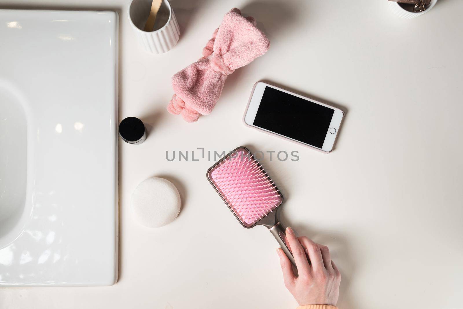 top view of bathroom accessories, toothbrush near hair brush, bow, mobile phone and face sponge on a white surface.