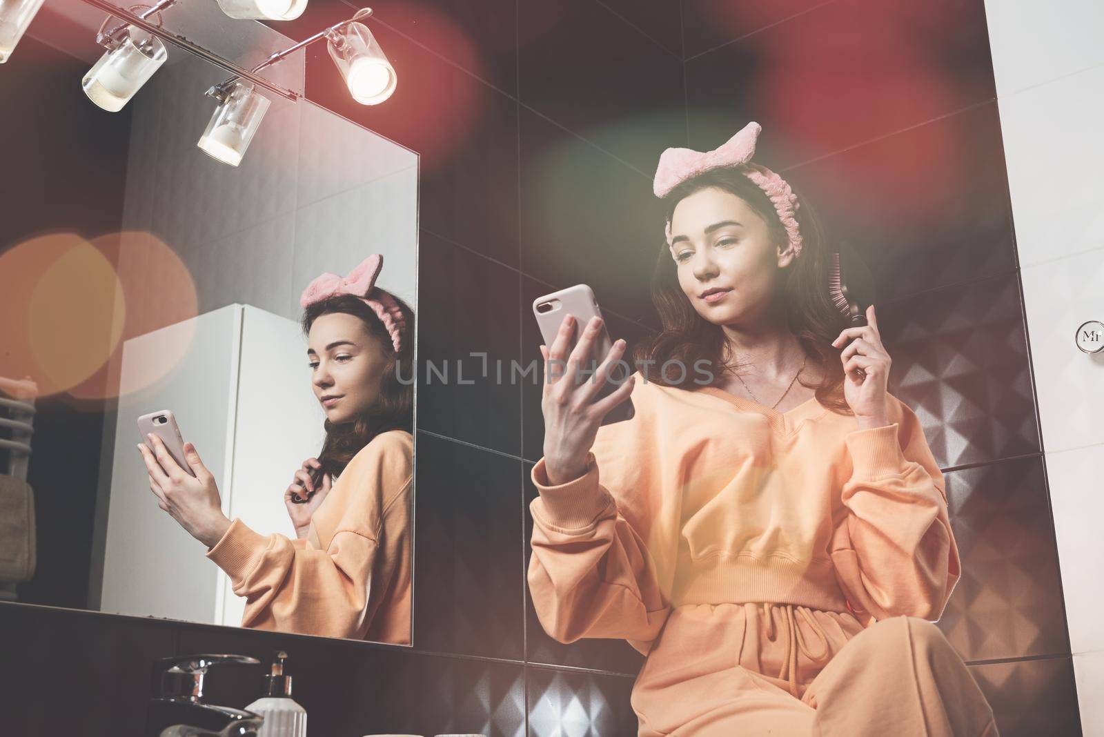 beautiful young woman communicating by the phone and doing her hair in a stylish bathroom. dressed in an orange sports suit and a bow by Ashtray25