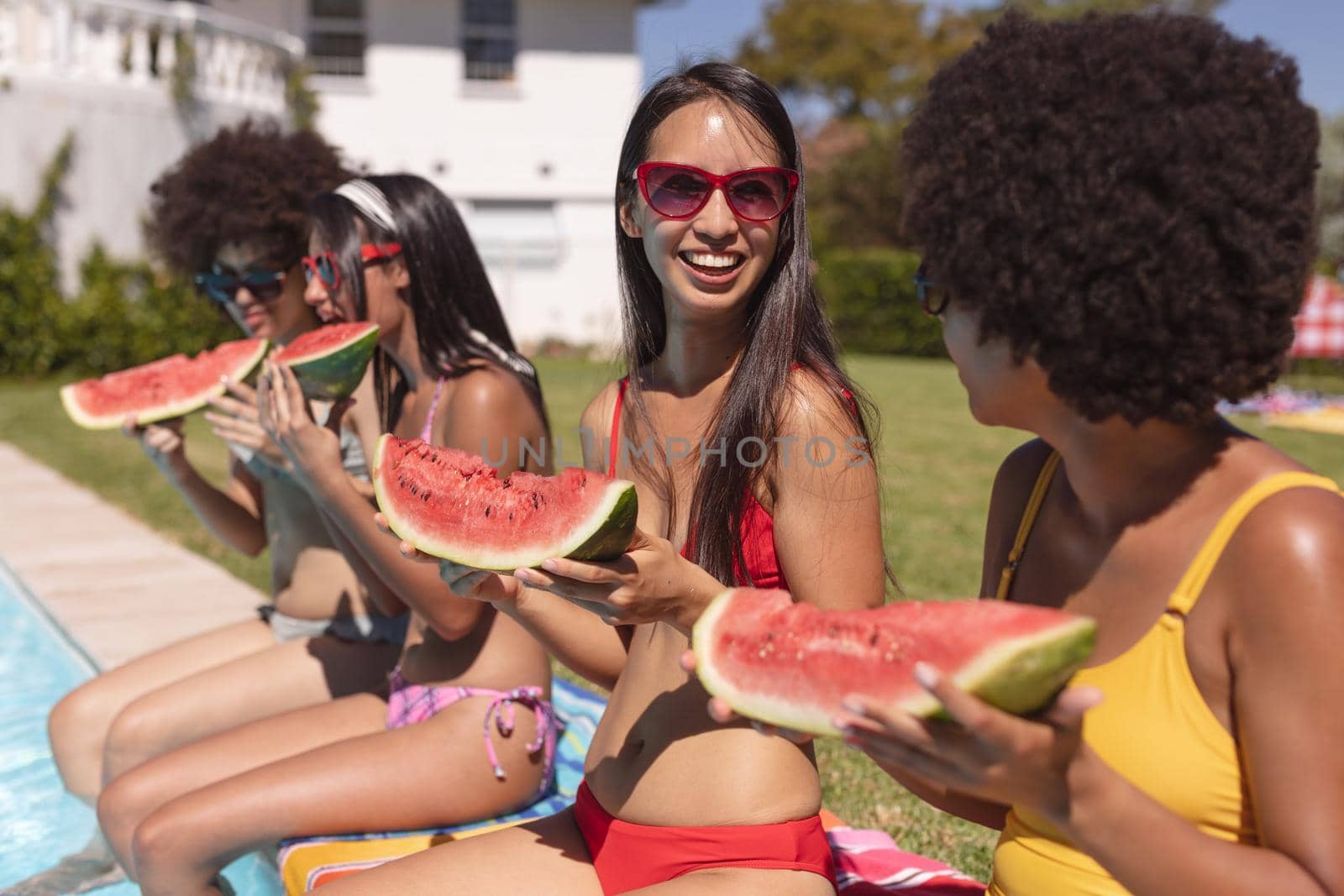 Diverse group of female friends eating watermelon sitting at the poolside talking by Wavebreakmedia