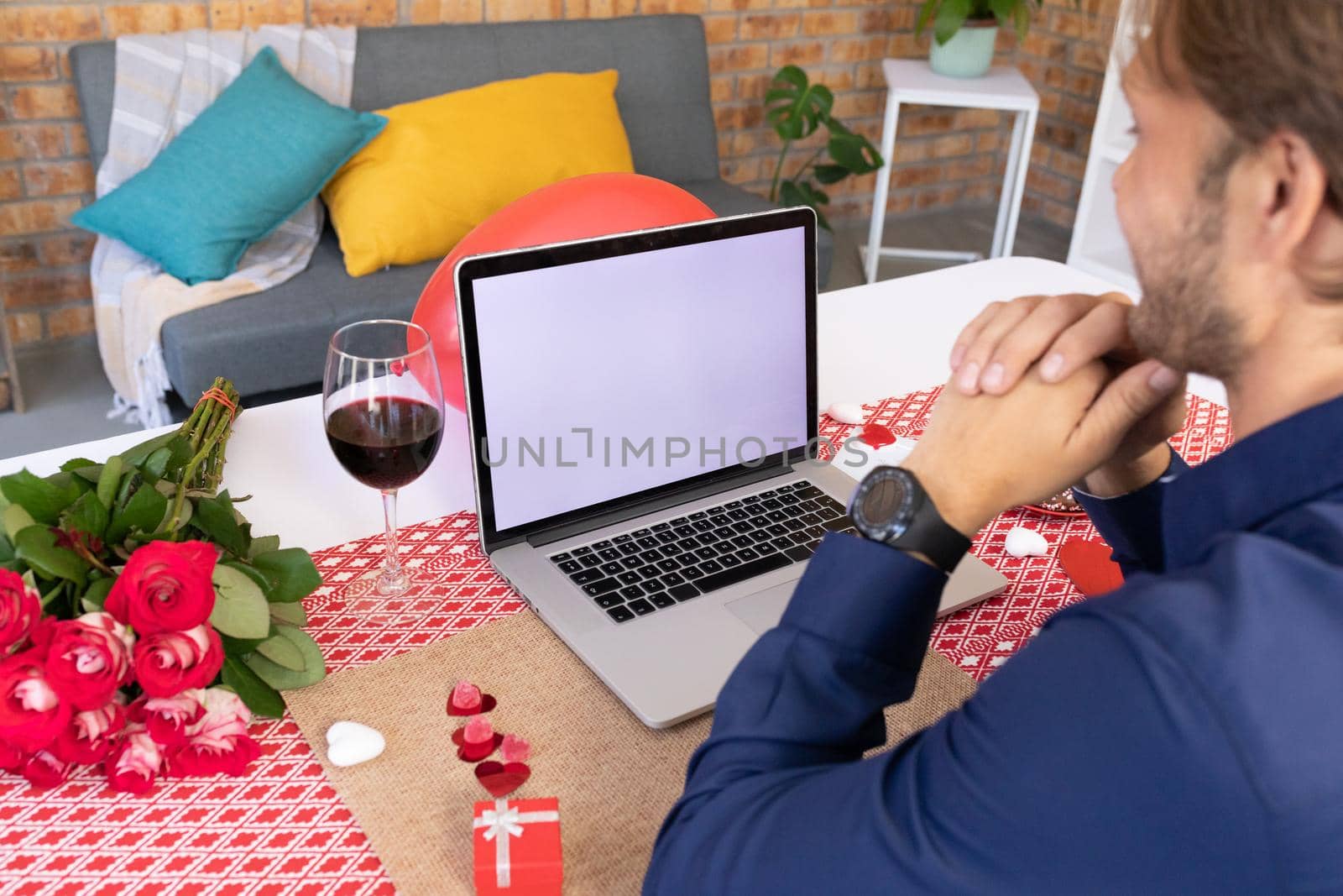 Caucasian man making video call using laptop with gift and glass of wine. online communication during quarantine lockdown