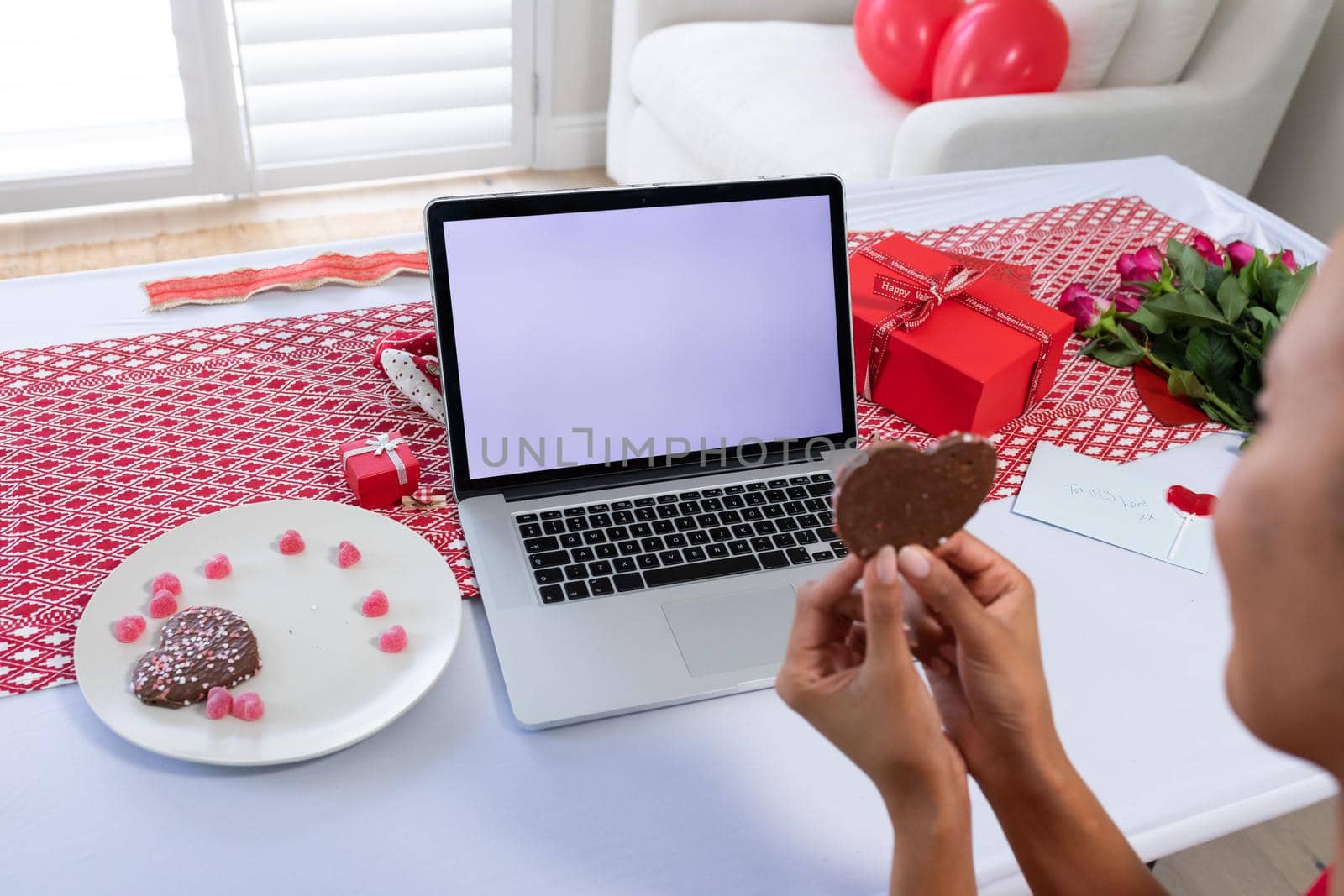 African american woman holding heart shaped cookie on videocall on laptop at home. distant celebration of valentines day online and dating concept