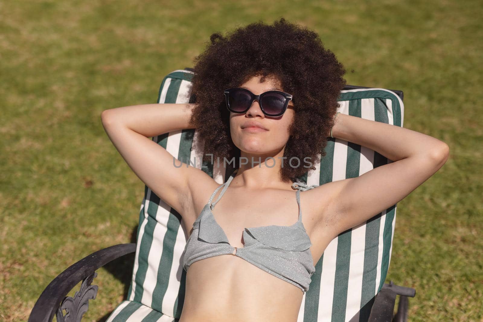 Mixed race woman wearing sunglasses sunbathing on sunbed in garden. Hanging out and relaxing outdoors in summer.