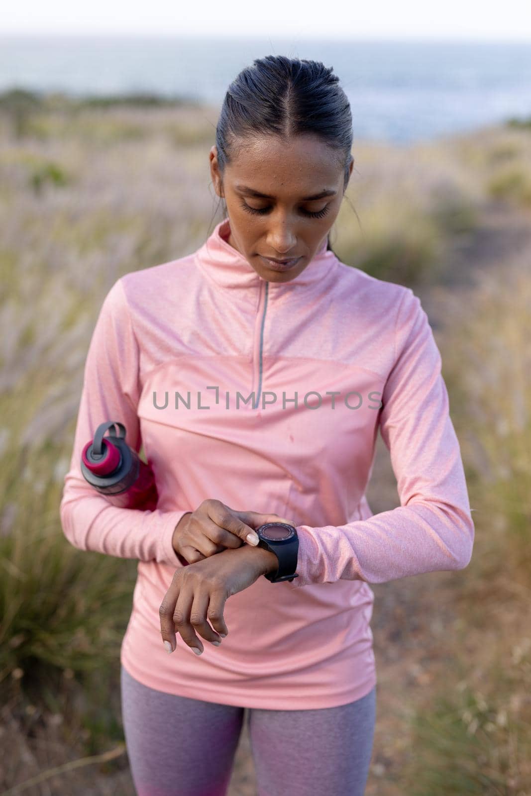 Portrait of fit african american woman in sportswear using smartwatch holding water in tall grass. healthy lifestyle, exercising in nature.