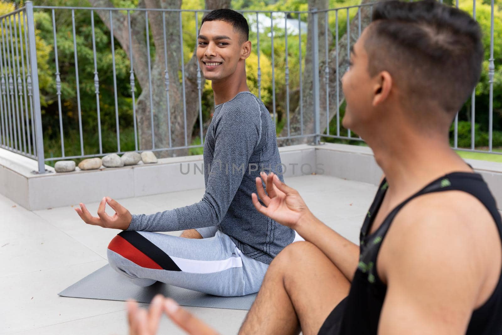Diverse gay male couple practicing yoga meditating and smiling on balcony. staying at home in isolation during quarantine lockdown.