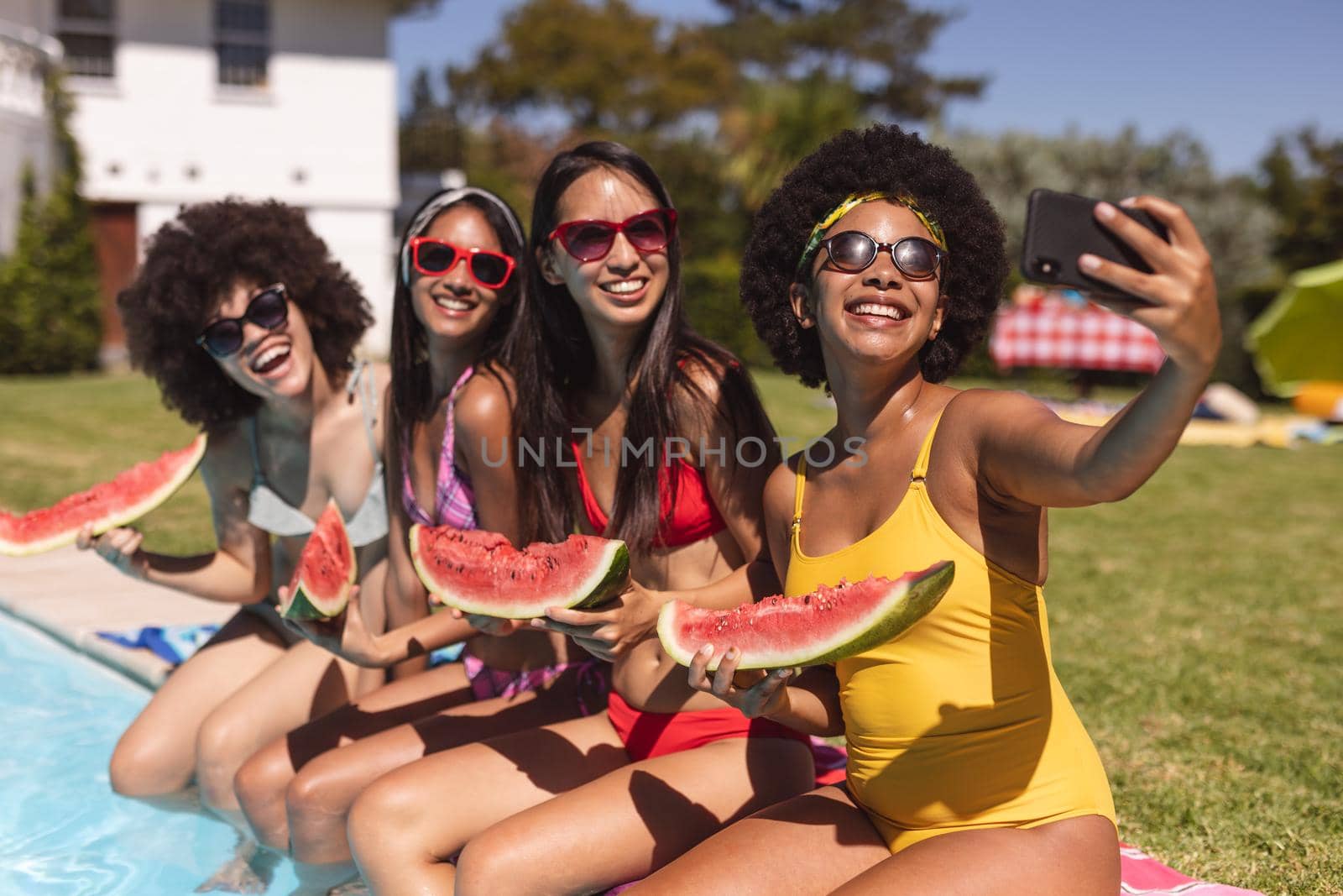 Diverse group of female friends taking selfie with watermelon sitting at the poolside. Hanging out and relaxing outdoors in summer.