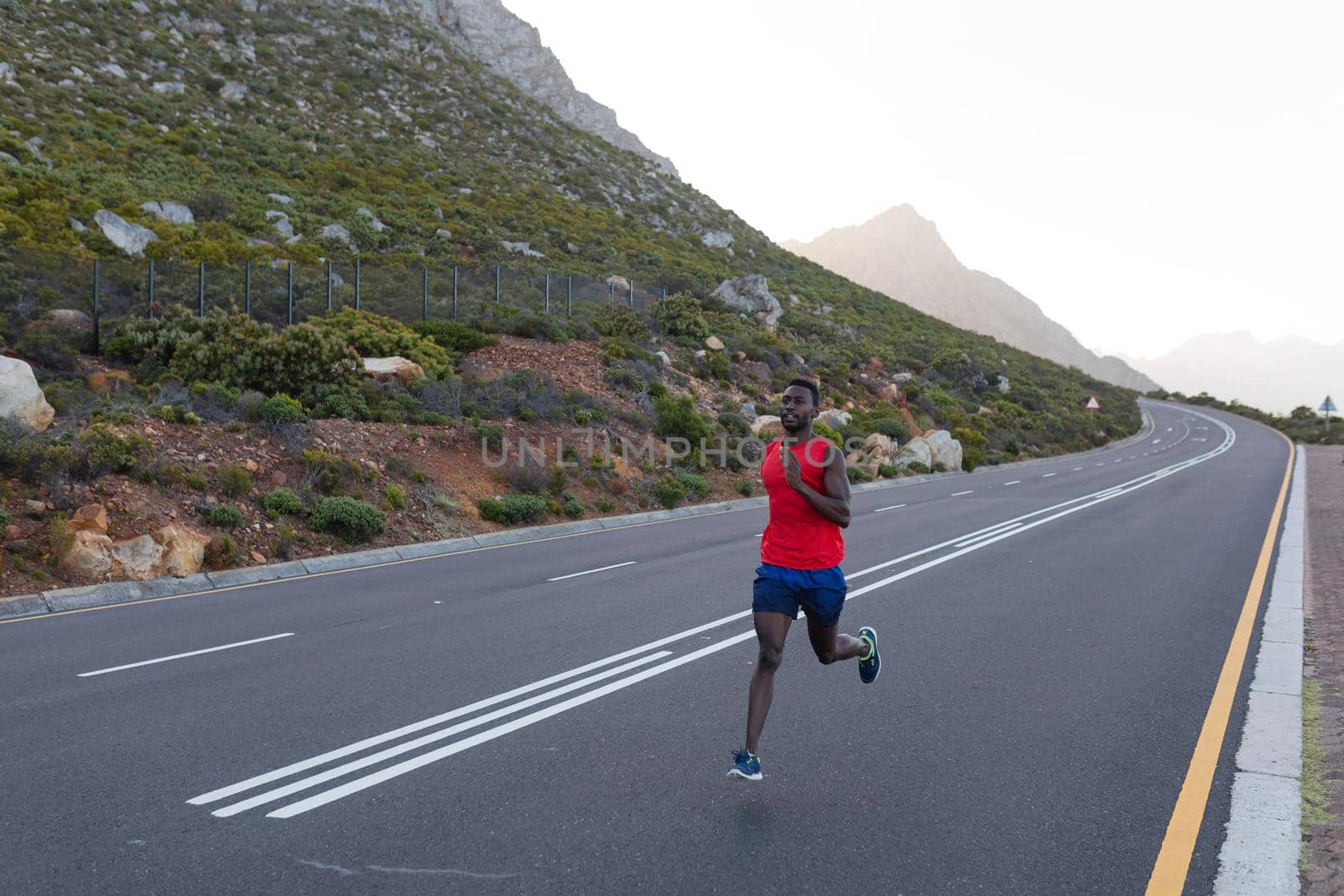 Fit african american man in sportswear running on a coastal road. looking at the camera and smiling. healthy lifestyle, exercising in nature.